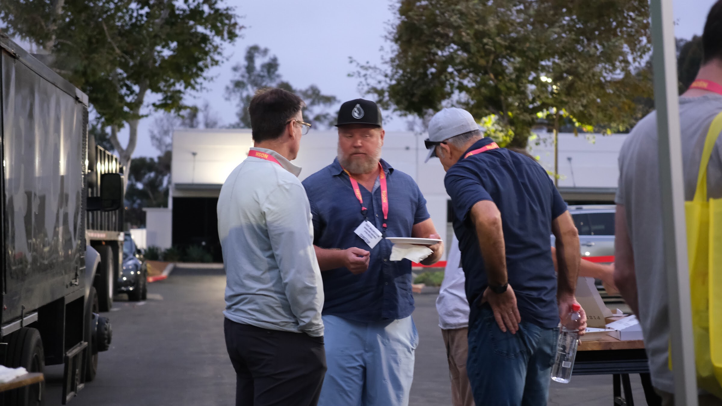Three men in lanyards talk beside a dark food truck at dusk, one holding a plate.