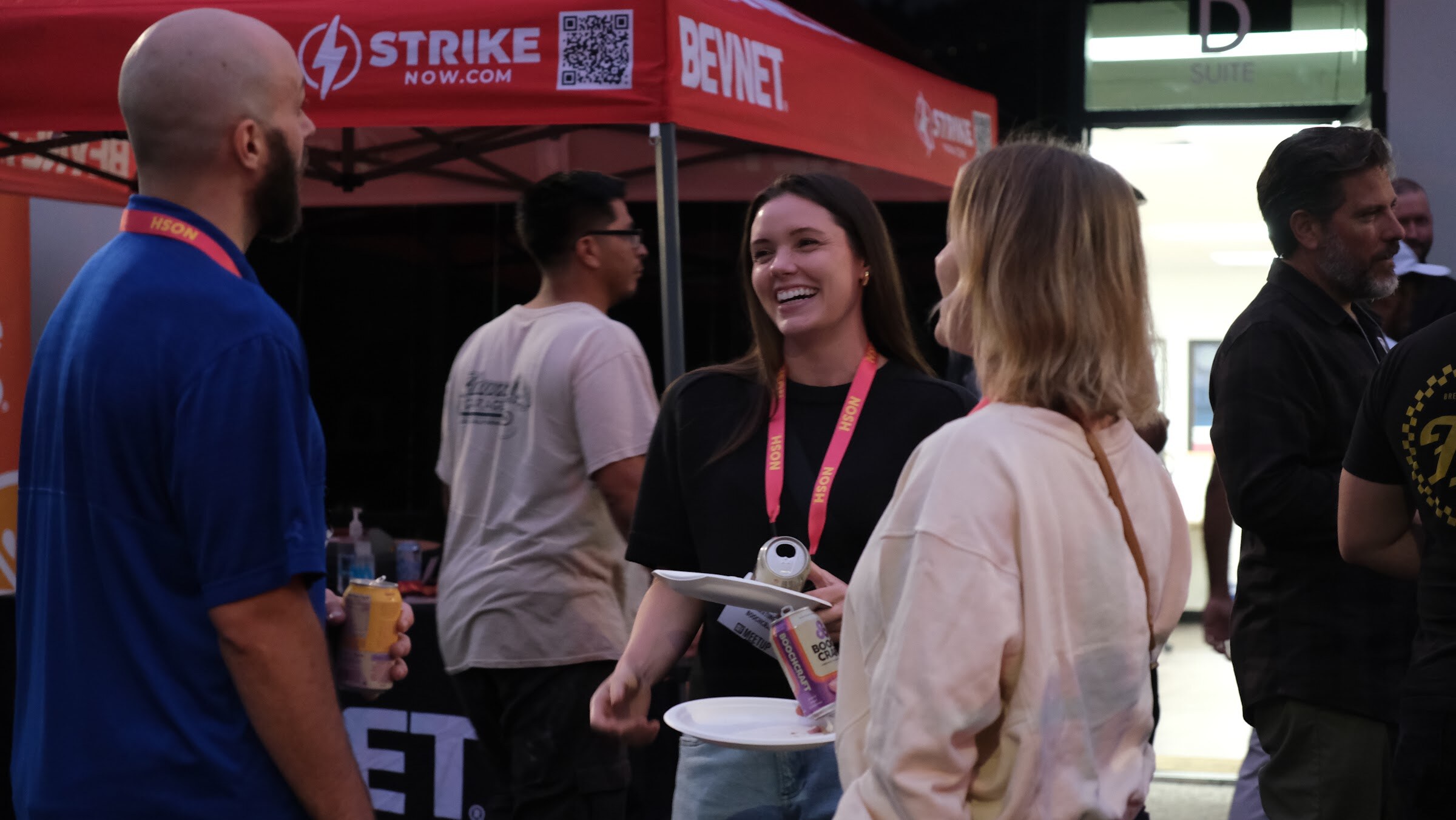 A smiling woman holding a can talks with another woman and a man in a blue shirt in front of a red BevNET tent.