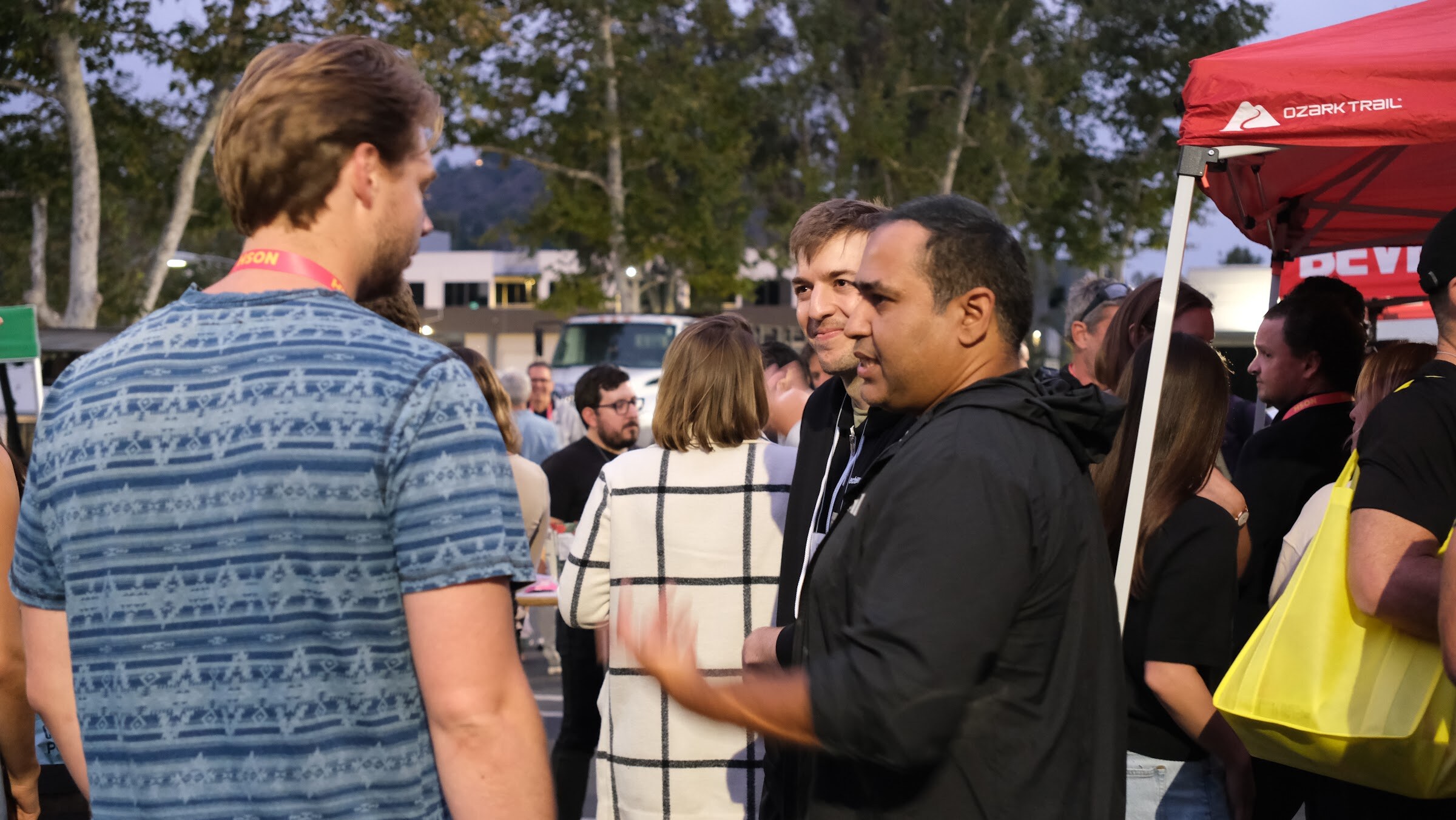 Two men in conversation in a crowd with a red tent and trees in the background.