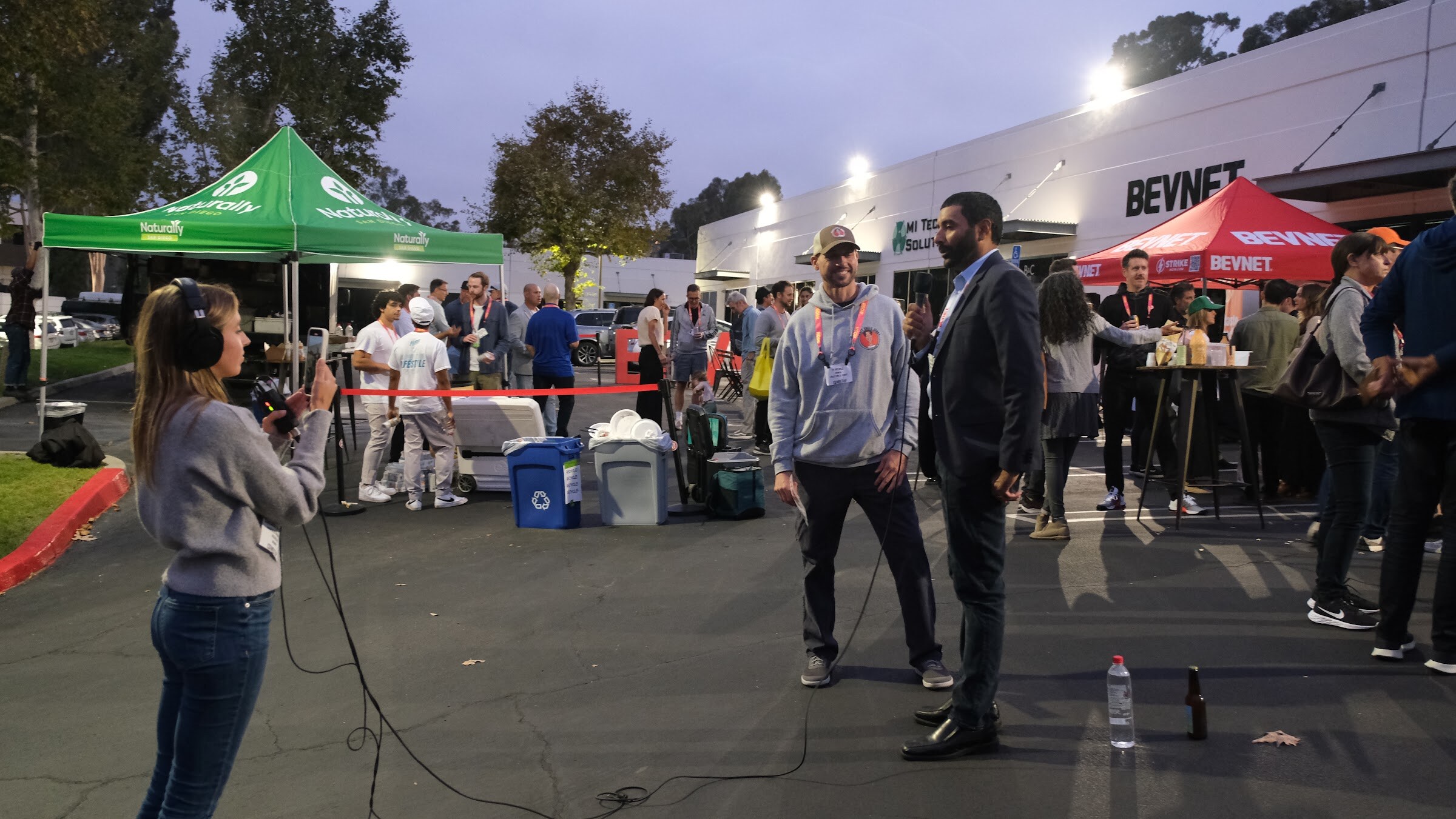 A man in a hooded sweatshirt is interviewed by a man in a blazer while a woman in headphones holds a camera on a tripod in a parking lot.