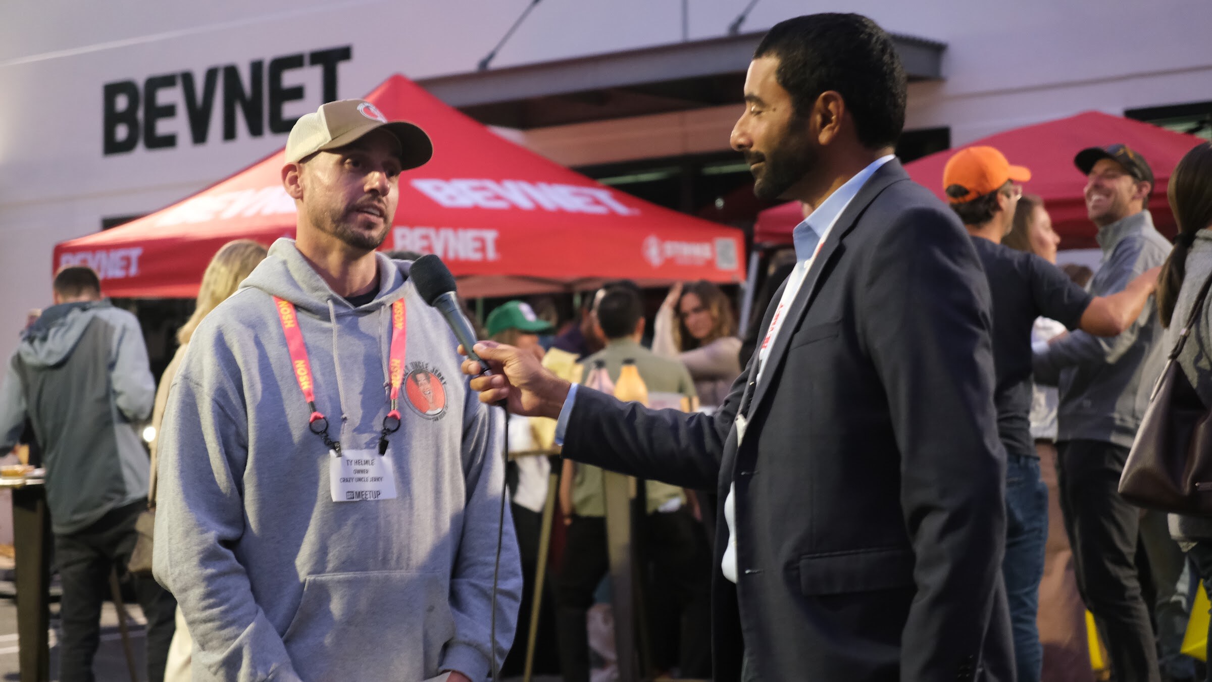 A man in a cap and grey hooded sweatshirt speaks into a handheld microphone held by a man in a dark blazer, with a BevNET tent behind them.