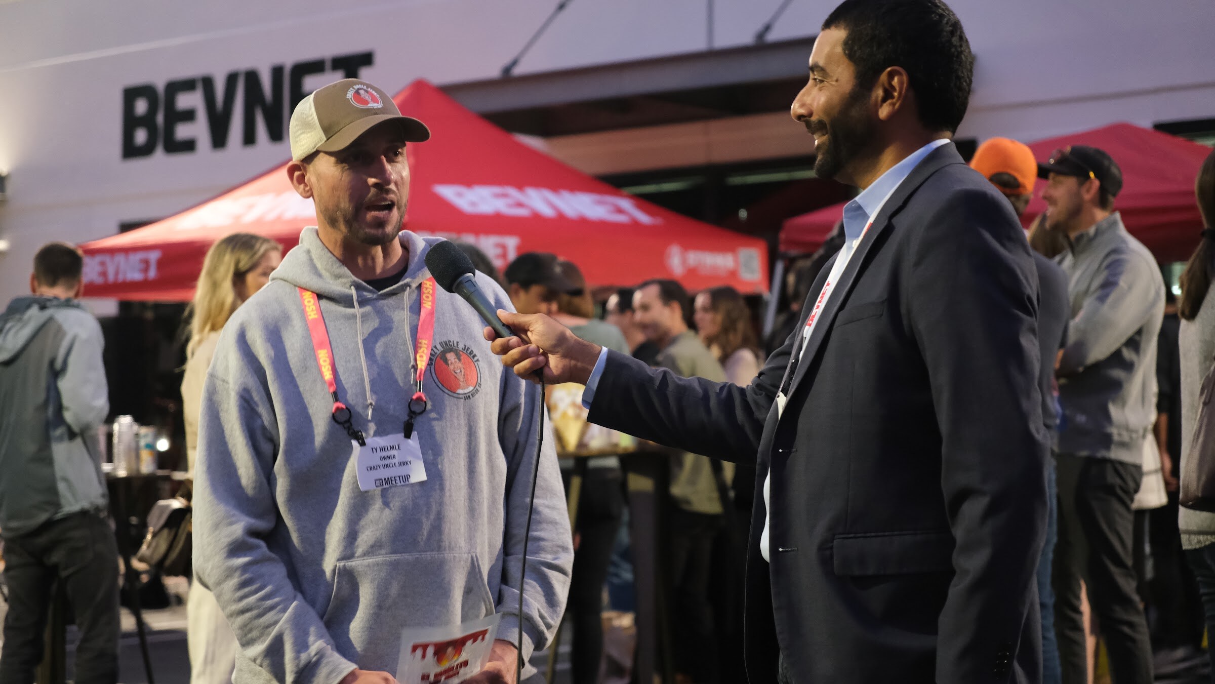 A man in a cap and grey sweatshirt answers a question from a man in a blazer in front of a BEVNET sign and red tent at dusk.