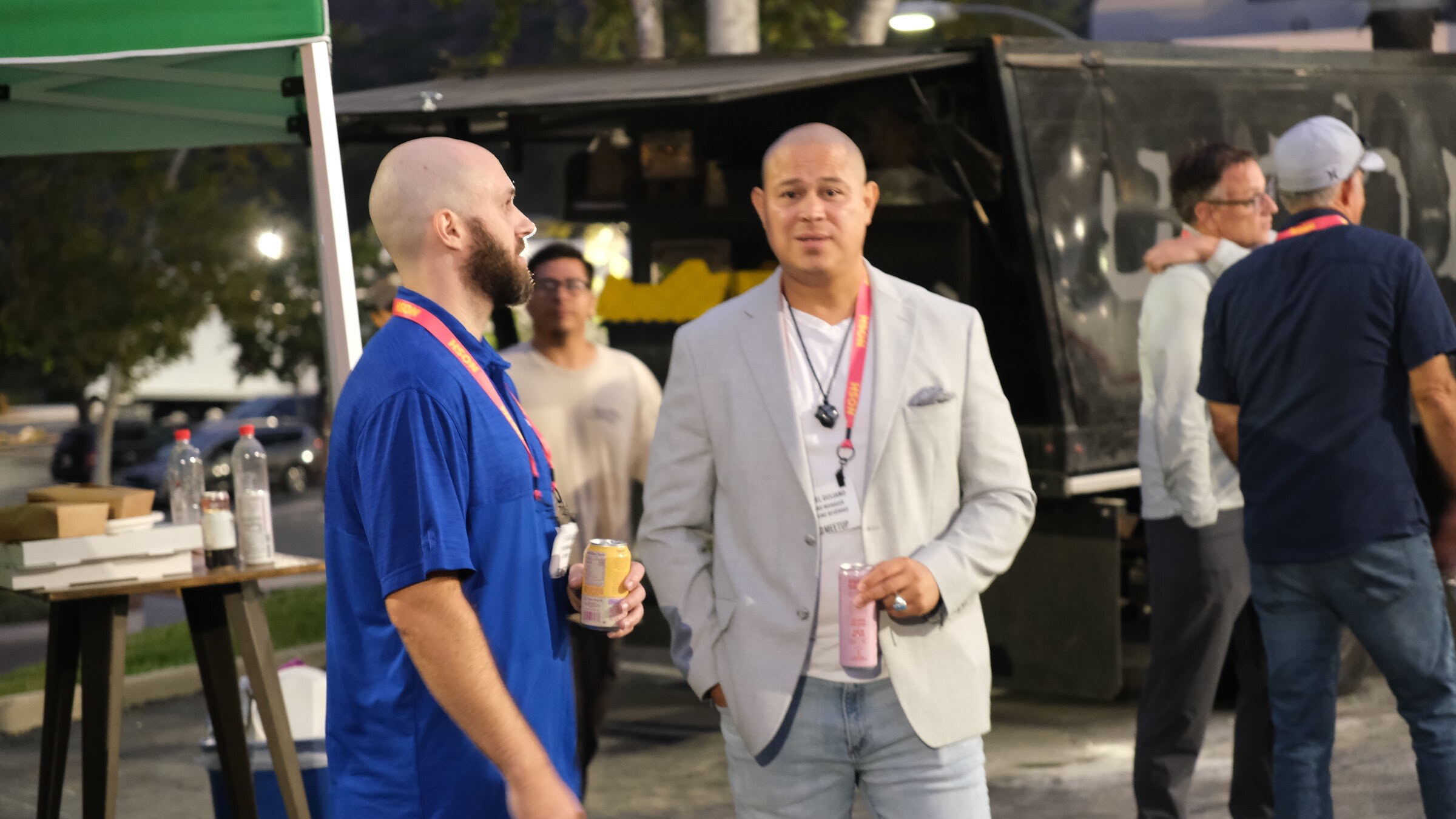A bald man in a blue polo and a man in a light grey blazer talk beside a food truck at night, each holding a drink.