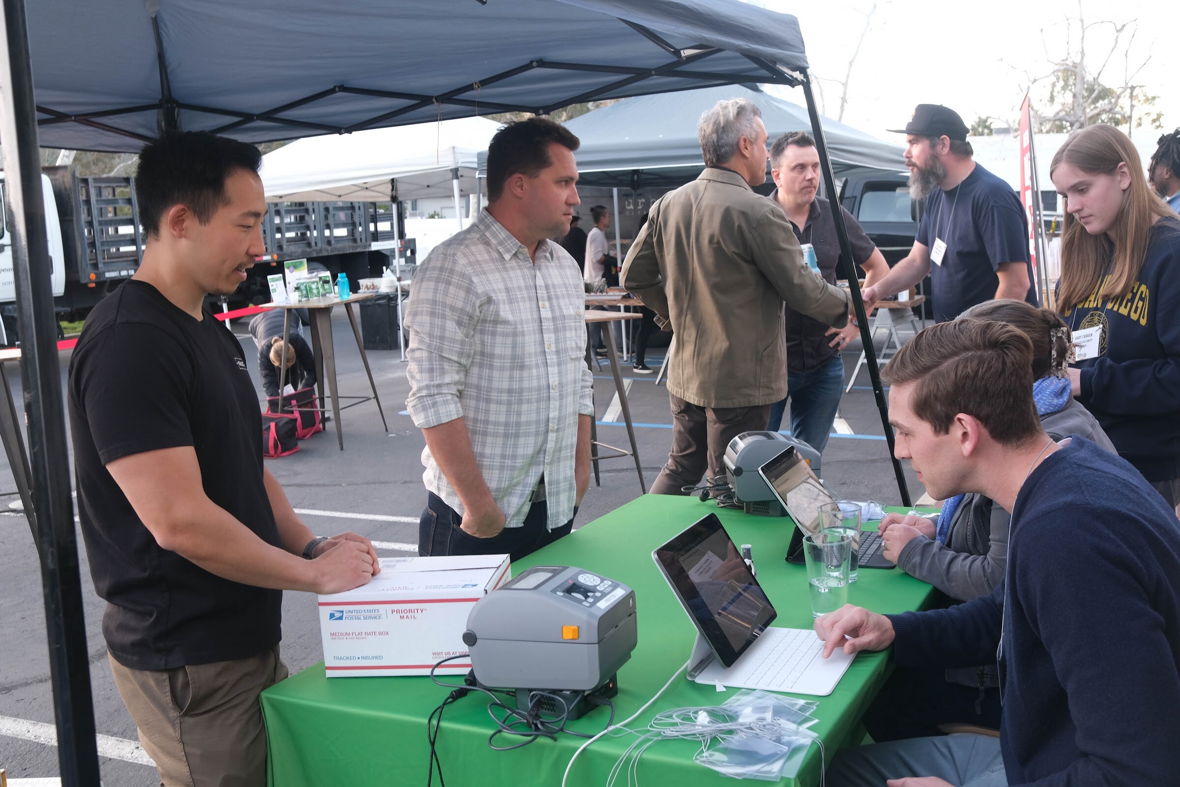 Several people stand and sit around a green-covered check-in table with a label printer, laptop and an envelope, under a dark canopy outdoors.