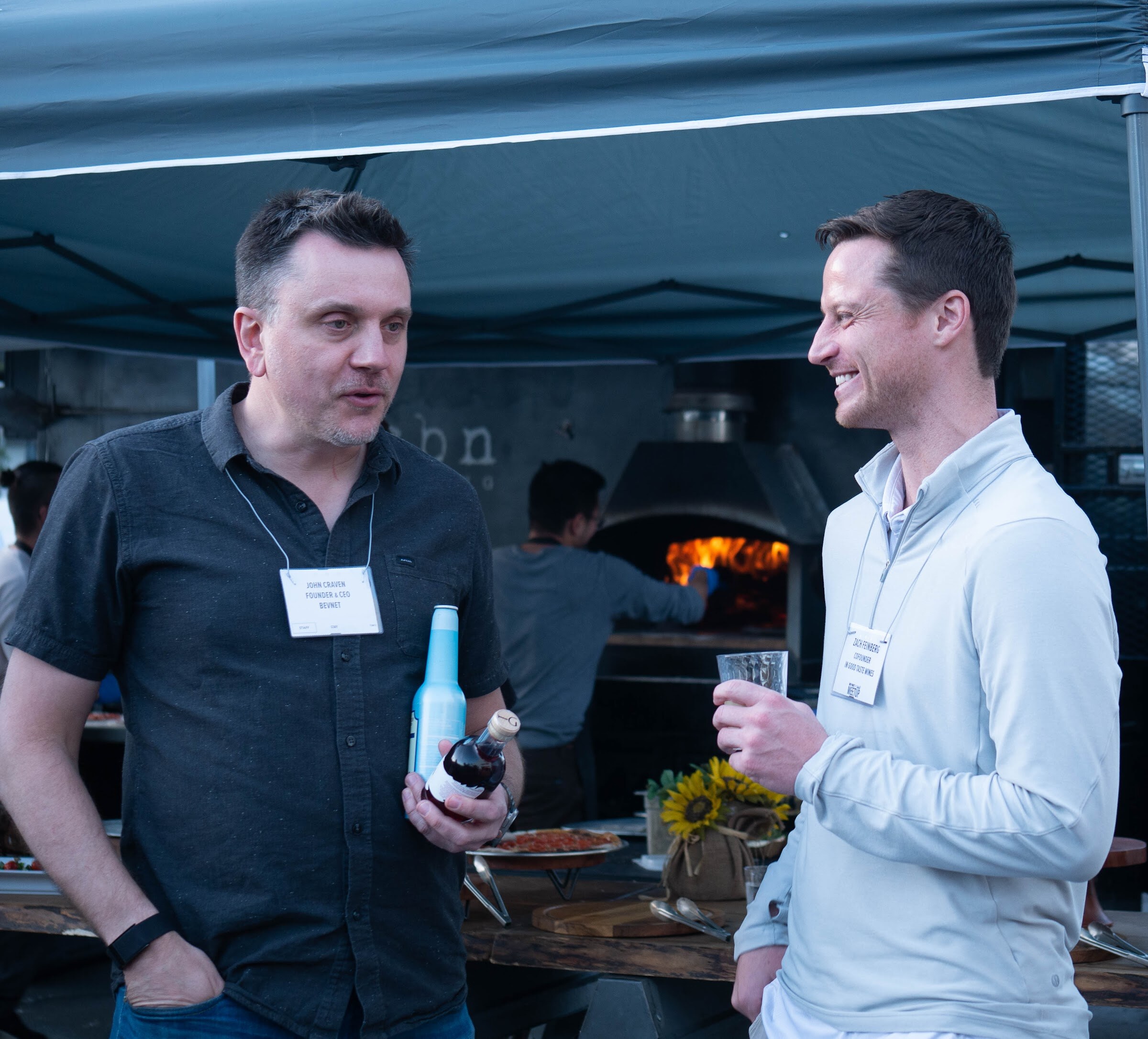 Two men wearing name badges talk while holding drinks, with a wood-fired pizza oven glowing behind them under a canopy.