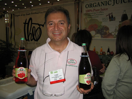 A smiling man with a name badge and an Elite Naturel logo shirt holds two bottles of red juice. A Theo Chocolate sign and an organic juice poster are behind him.