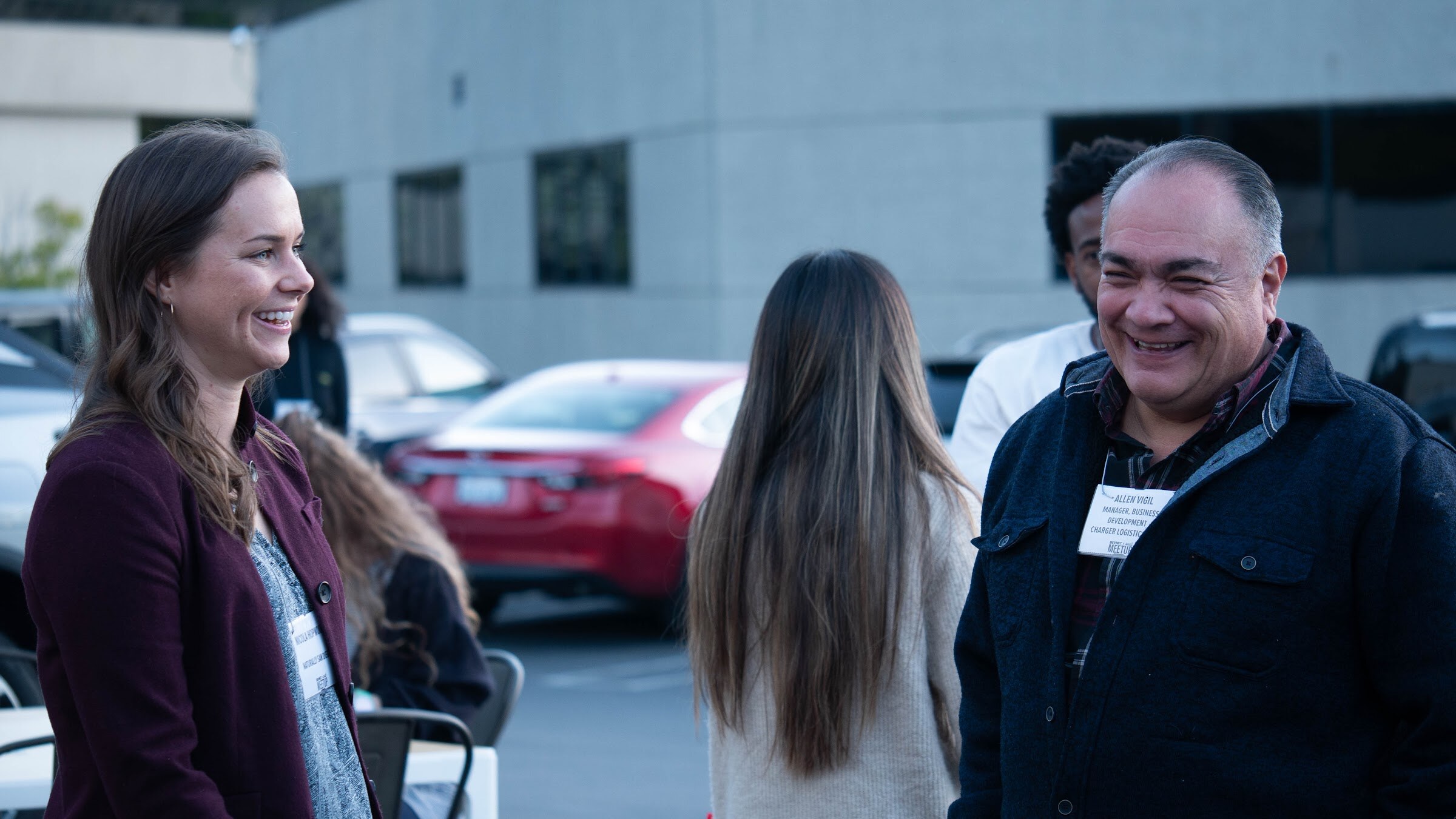 A smiling woman in a maroon jacket and a man in a dark jacket talk with others in a parking lot.
