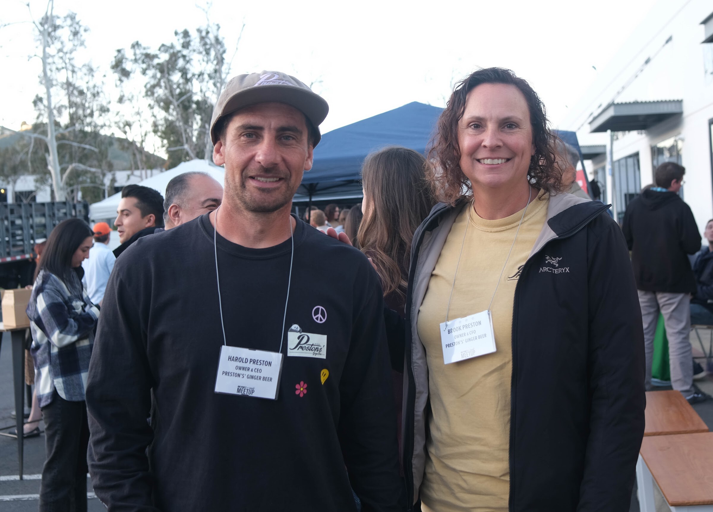 A man in a cap and a woman in a tan jacket, both wearing name badges on lanyards, smile for the camera outdoors at dusk.