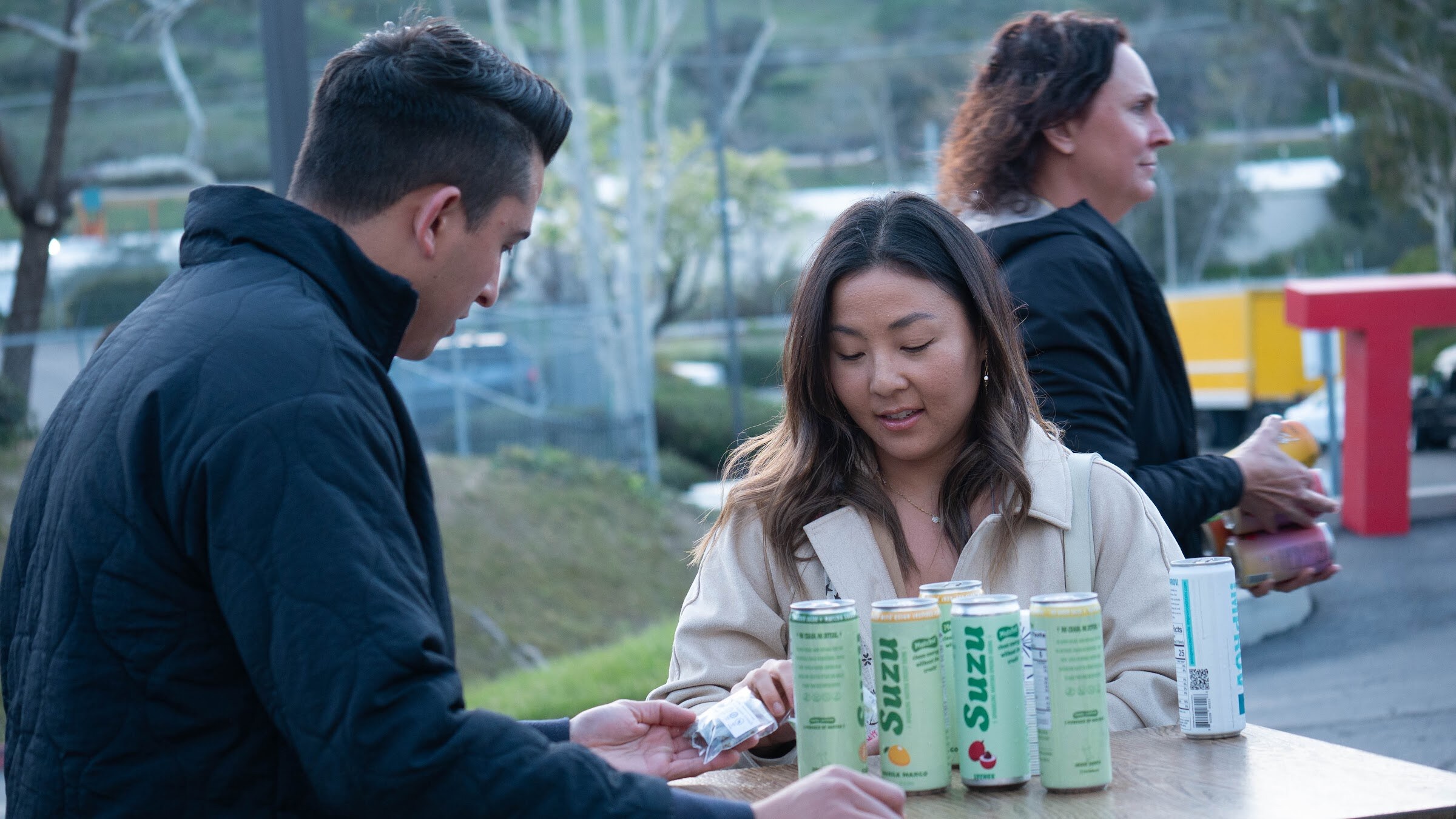 A man in a navy jacket and a woman in a cream coat sample cans of Suzu drinks at a table outdoors.
