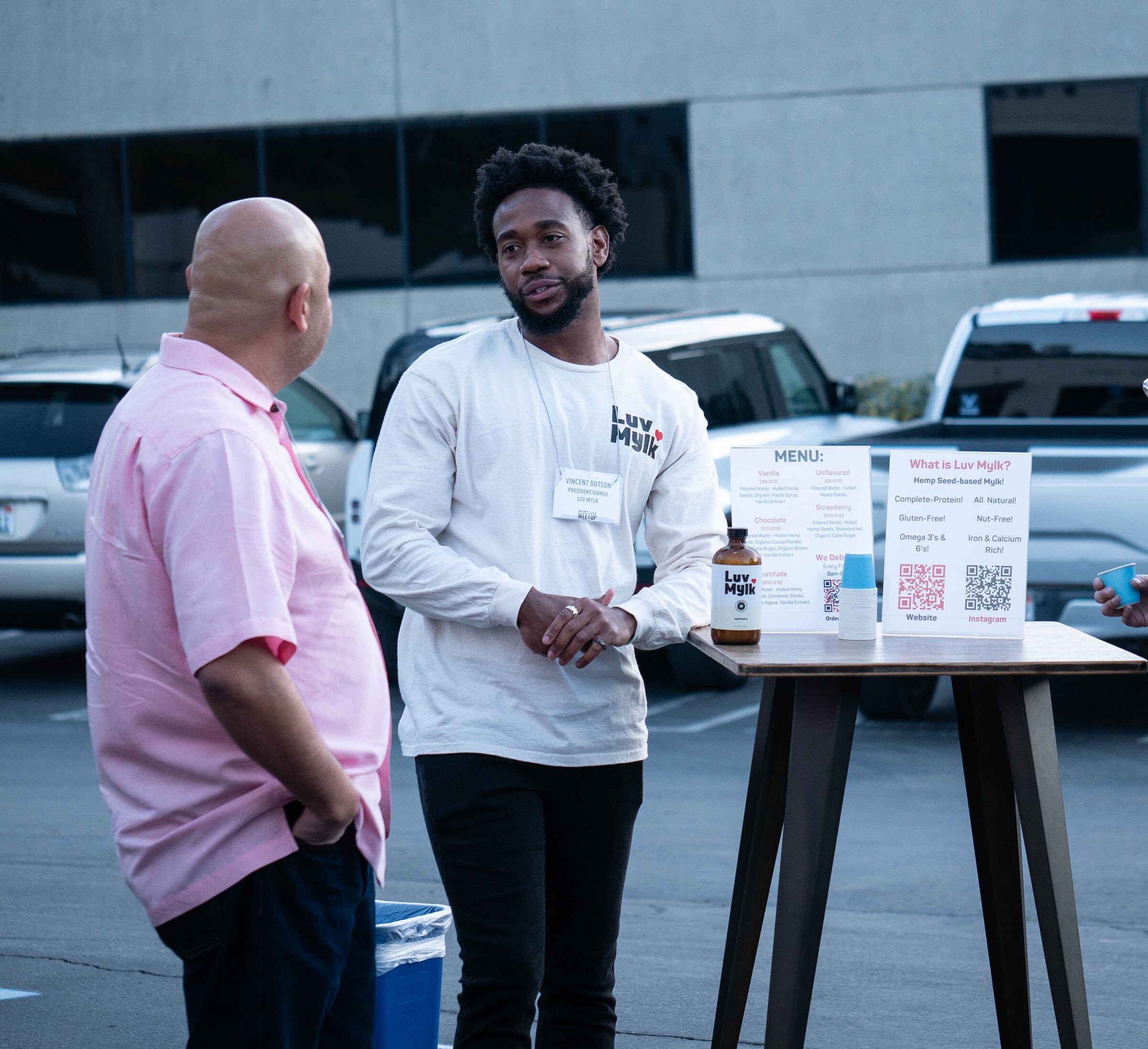 A man in a Luv Mylk shirt stands at a high-top table with a menu and QR code while another man in a pink shirt talks with him.