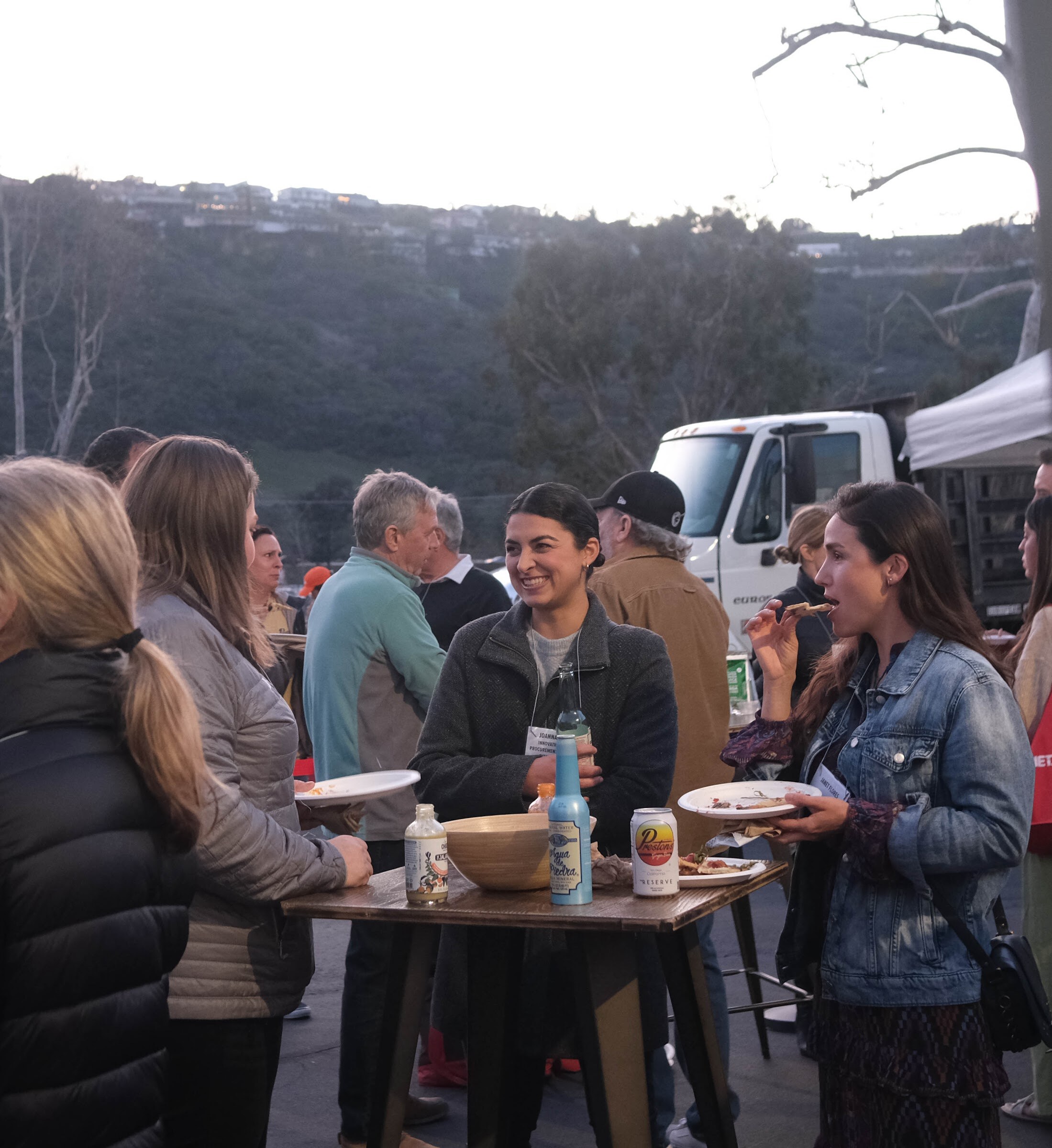 A group of people eat and talk around a small high-top table holding food and cans, with hills in the background.