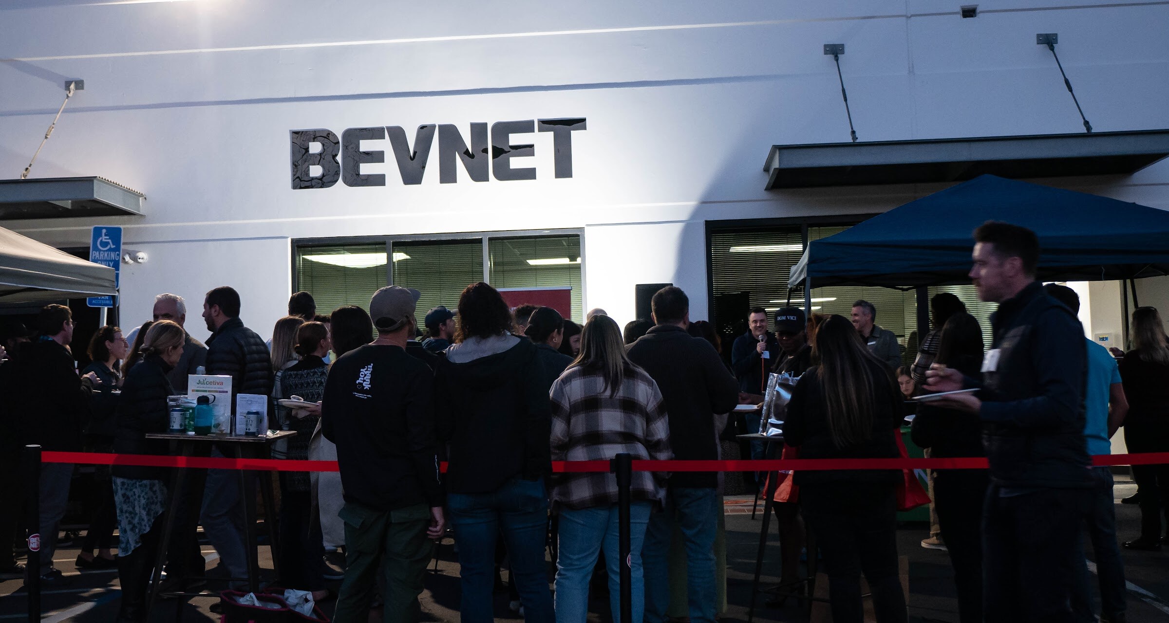 Wide shot of dozens of guests in front of a white building with a large BEVNET sign, with a red rope and tables in the foreground.