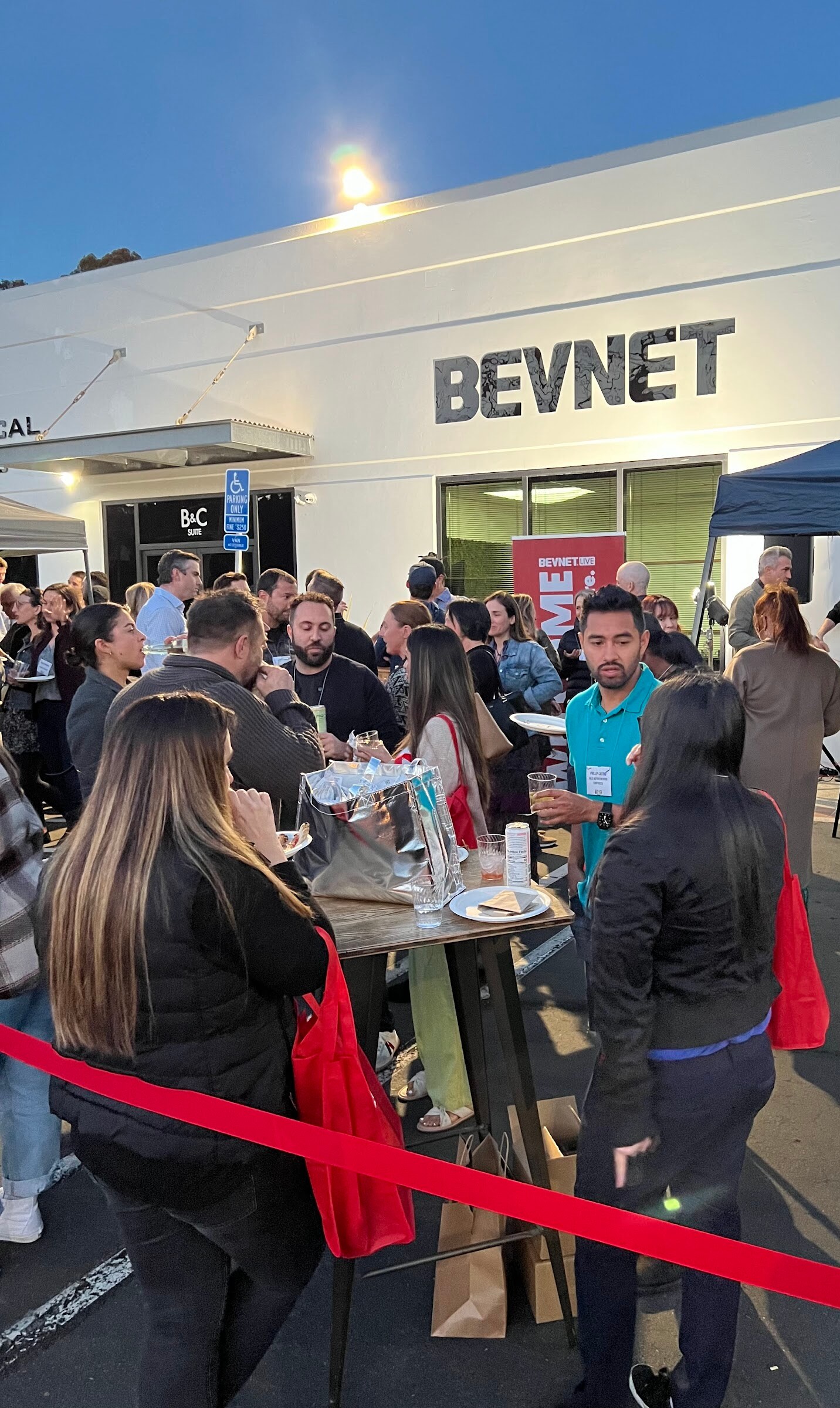 Evening shot of a crowd gathered in a parking lot in front of a white building with a BEVNET sign, with a red rope line in the foreground.