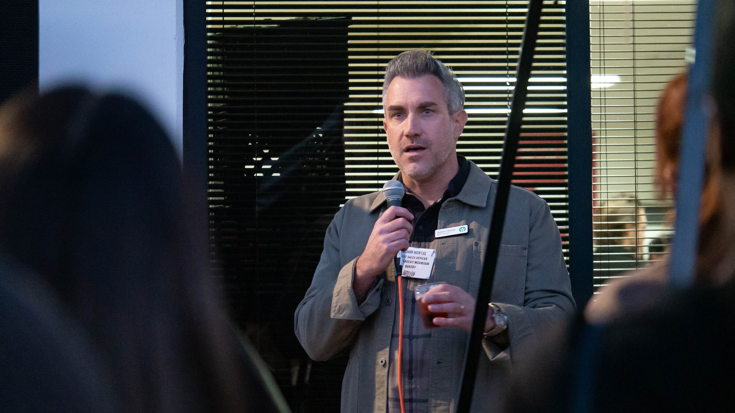 A man in an olive jacket and name badge speaks into a microphone inside the doorway of the office.