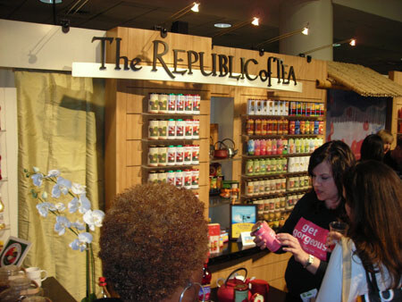 Two women look at products at The Republic of Tea booth. Wooden shelves hold rows of tea tins, and one woman holds a pink package marked get gorgeous. A white orchid sits at left.