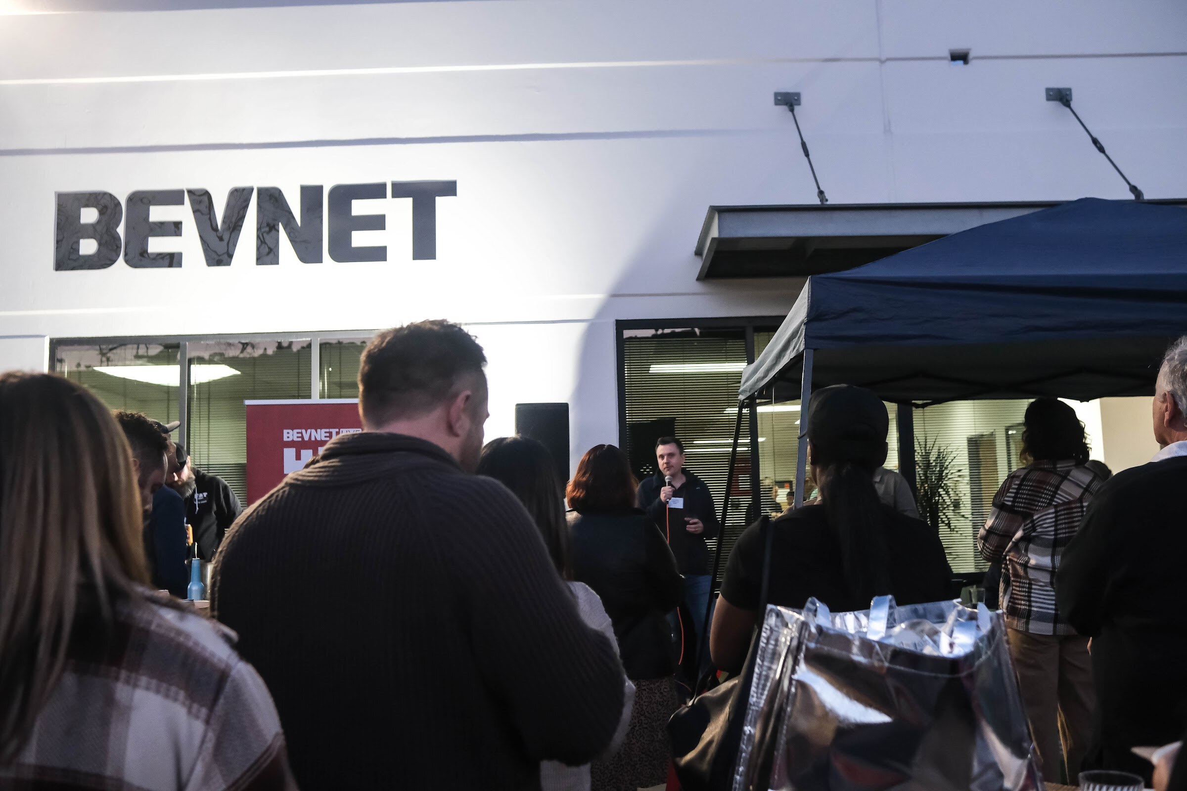 Crowd of guests in the foreground facing a white building with a large BEVNET sign and a dark blue canopy.