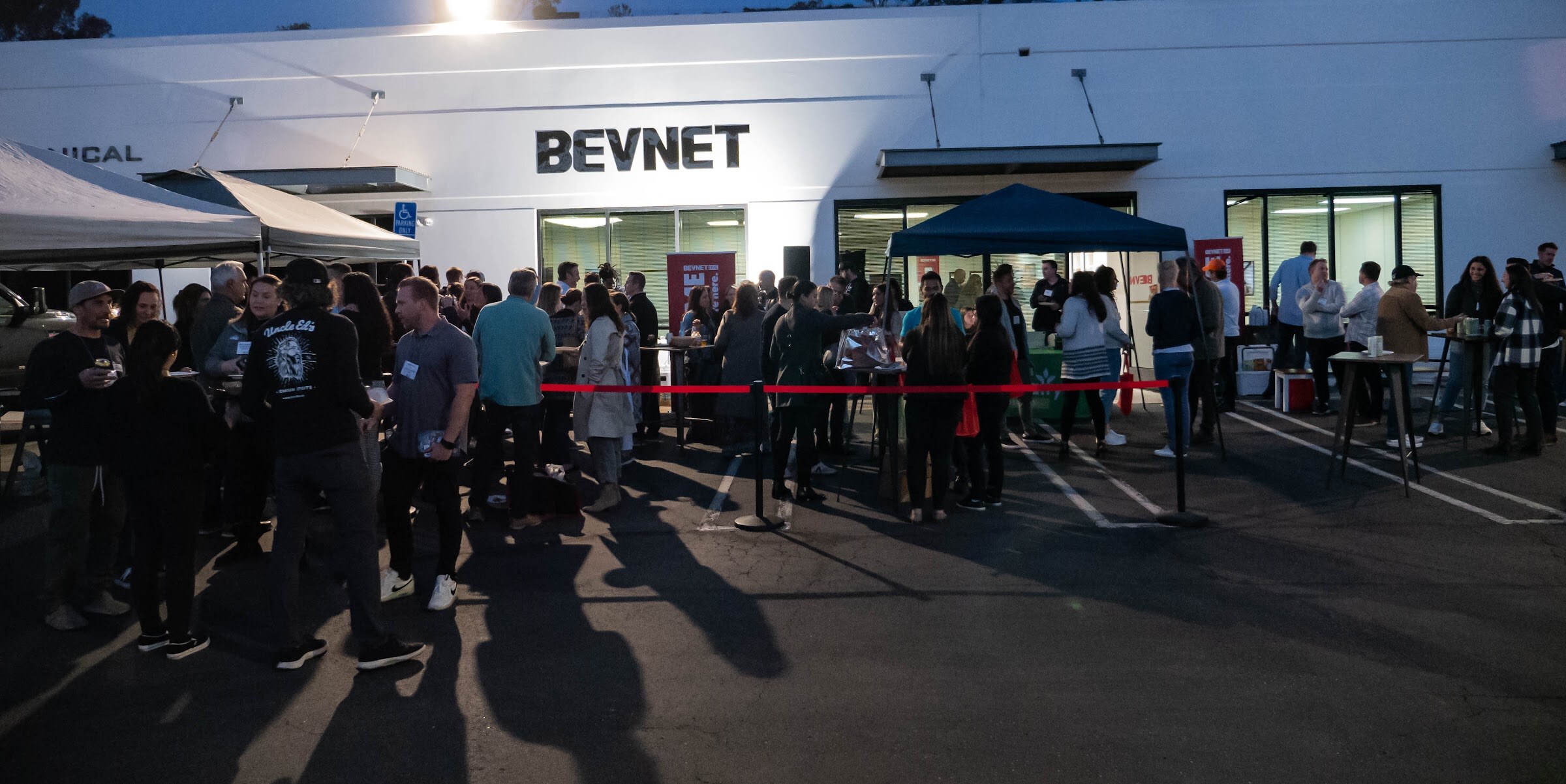 Wide view of many people gathered in front of the BevNET building with tents and a red rope line at dusk.