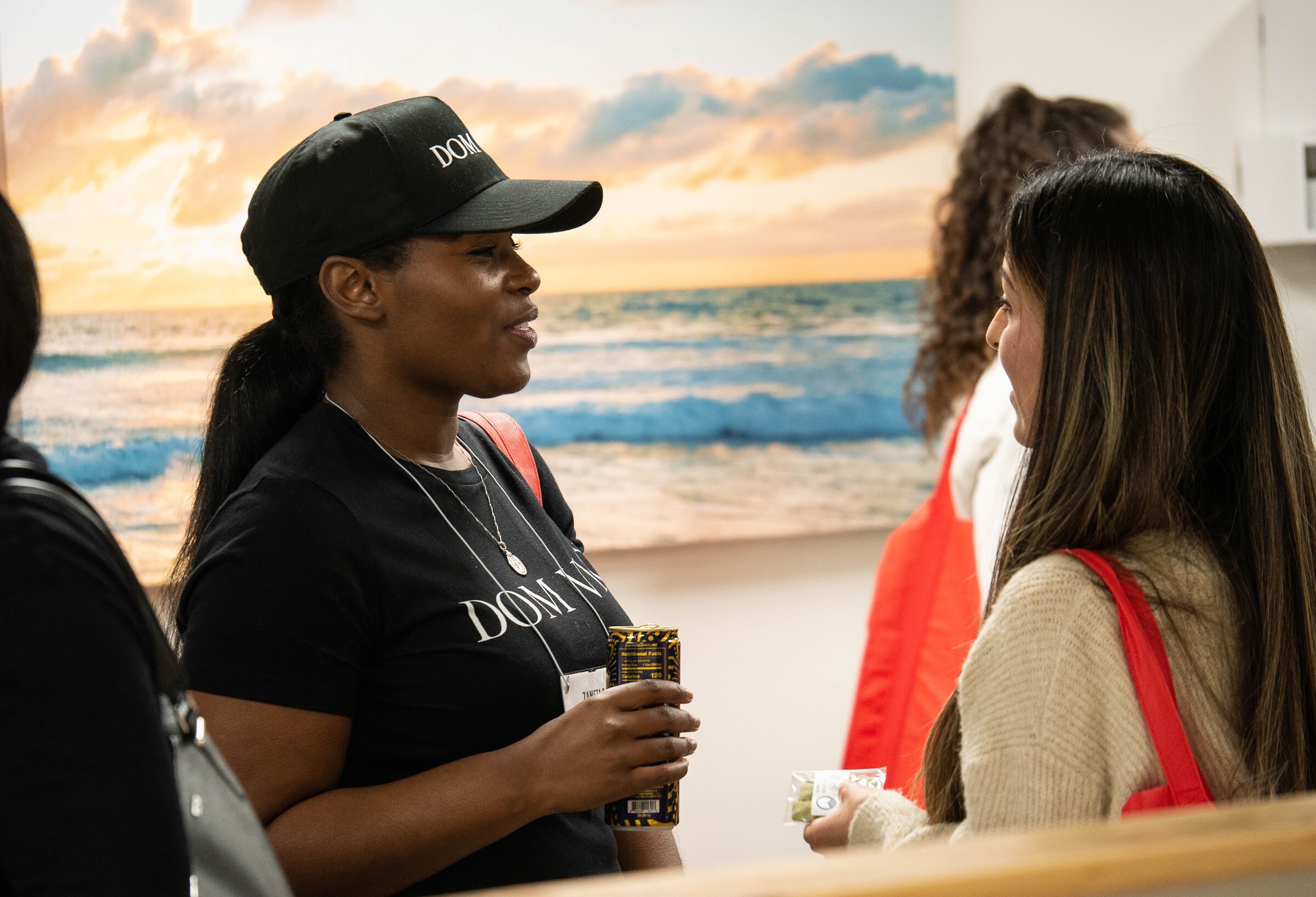 A woman in a black Down Good cap talks with another woman in front of a large wall photo of ocean waves at sunset.