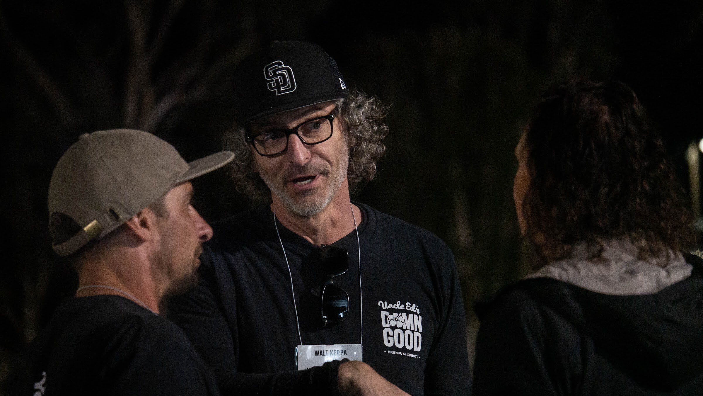 A man in a black Uncle Ed's Down Good shirt and glasses talks with two other guests outdoors at night.