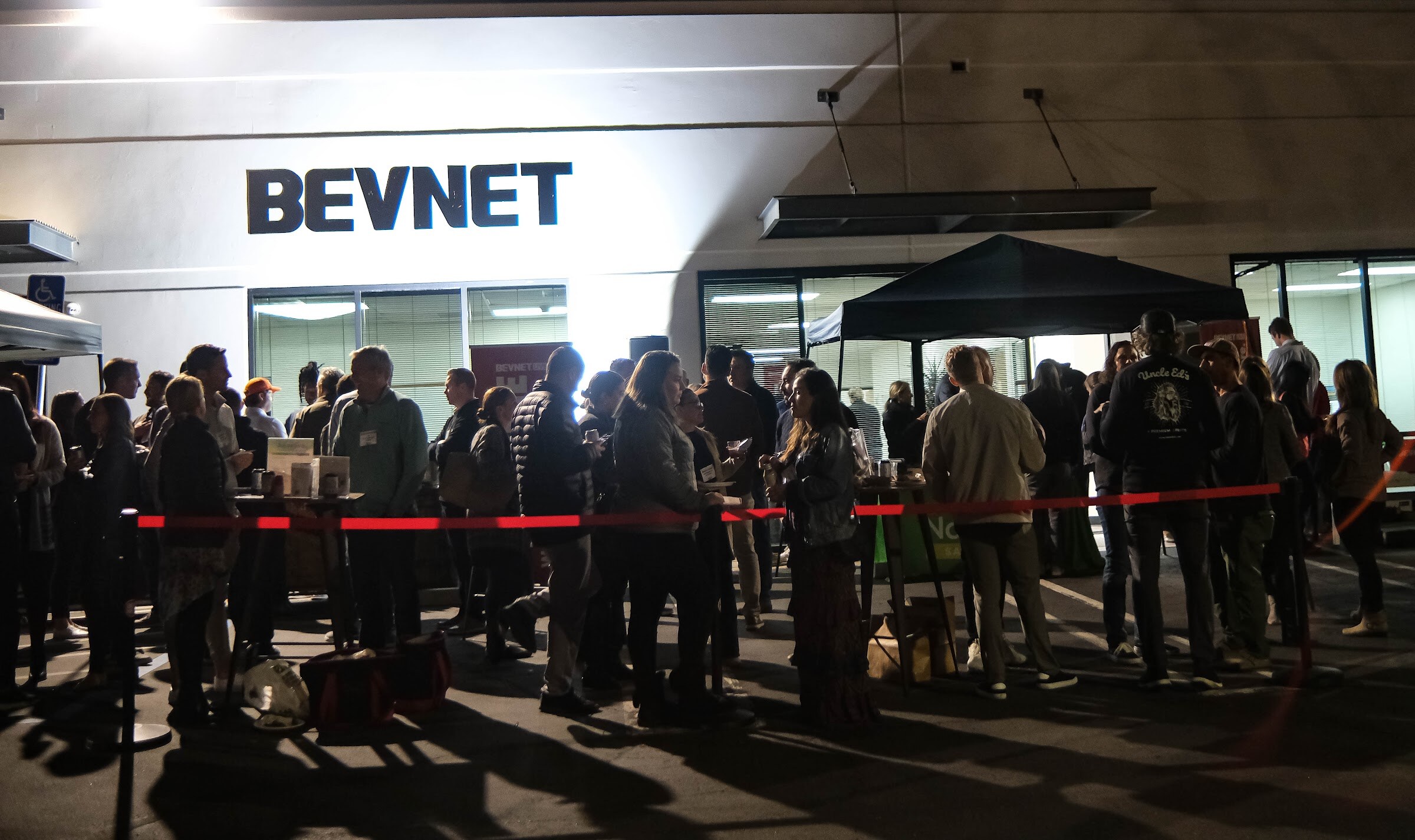 Night view of a crowd of people standing behind a red rope in front of a white building with the BEVNET sign.
