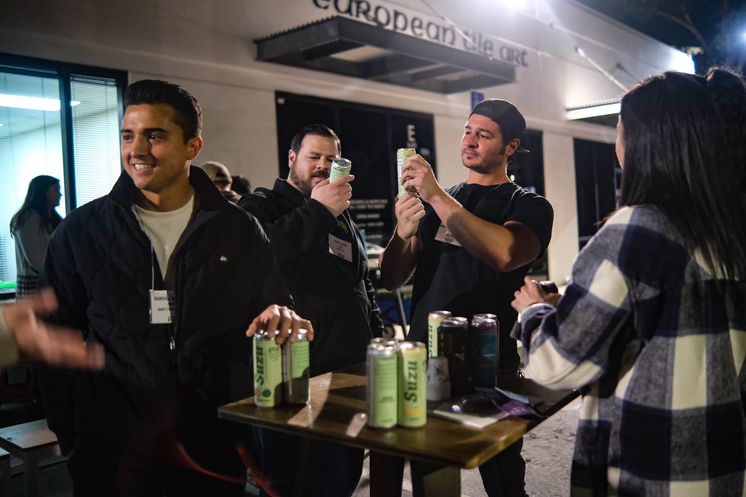 Three men at a wooden table lift and examine green and white cans of Suzu drinks while a woman in a plaid coat looks on.