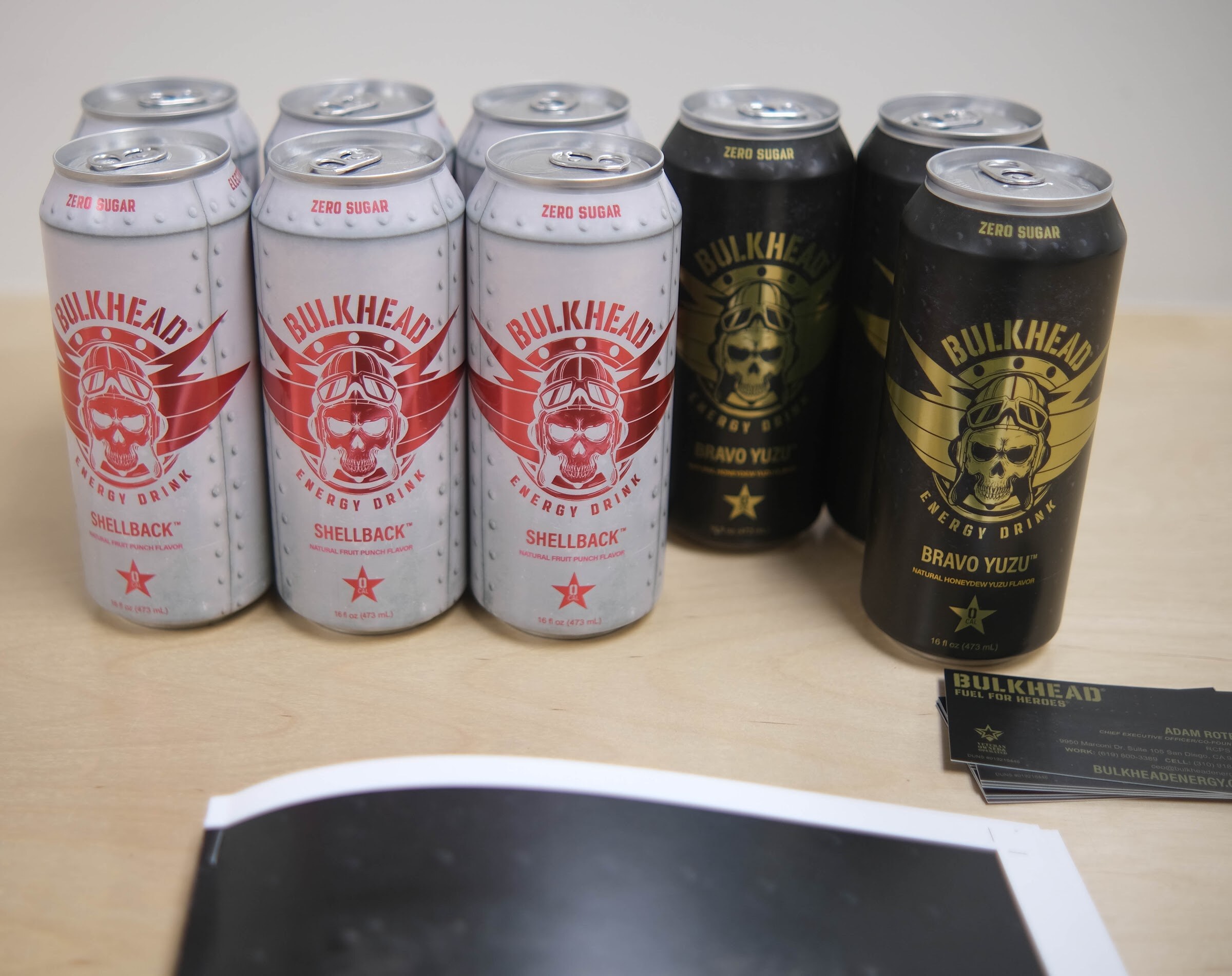 Six cans of Bulkhead zero sugar energy drink in red-silver and black-gold designs on a light surface with business cards.