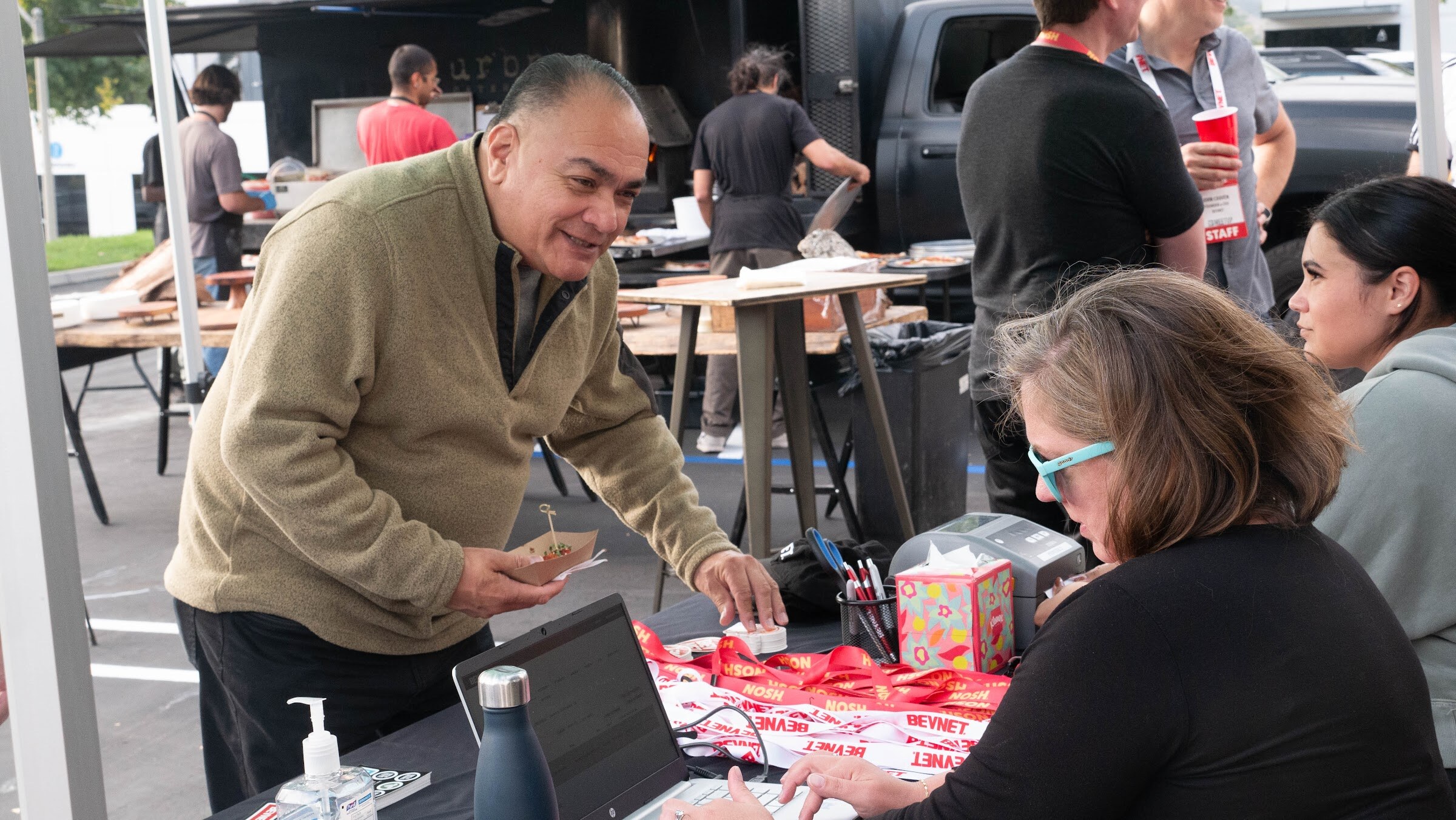 A man in a tan sweater smiles at a woman at a laptop on the registration table with lanyards and a small display.