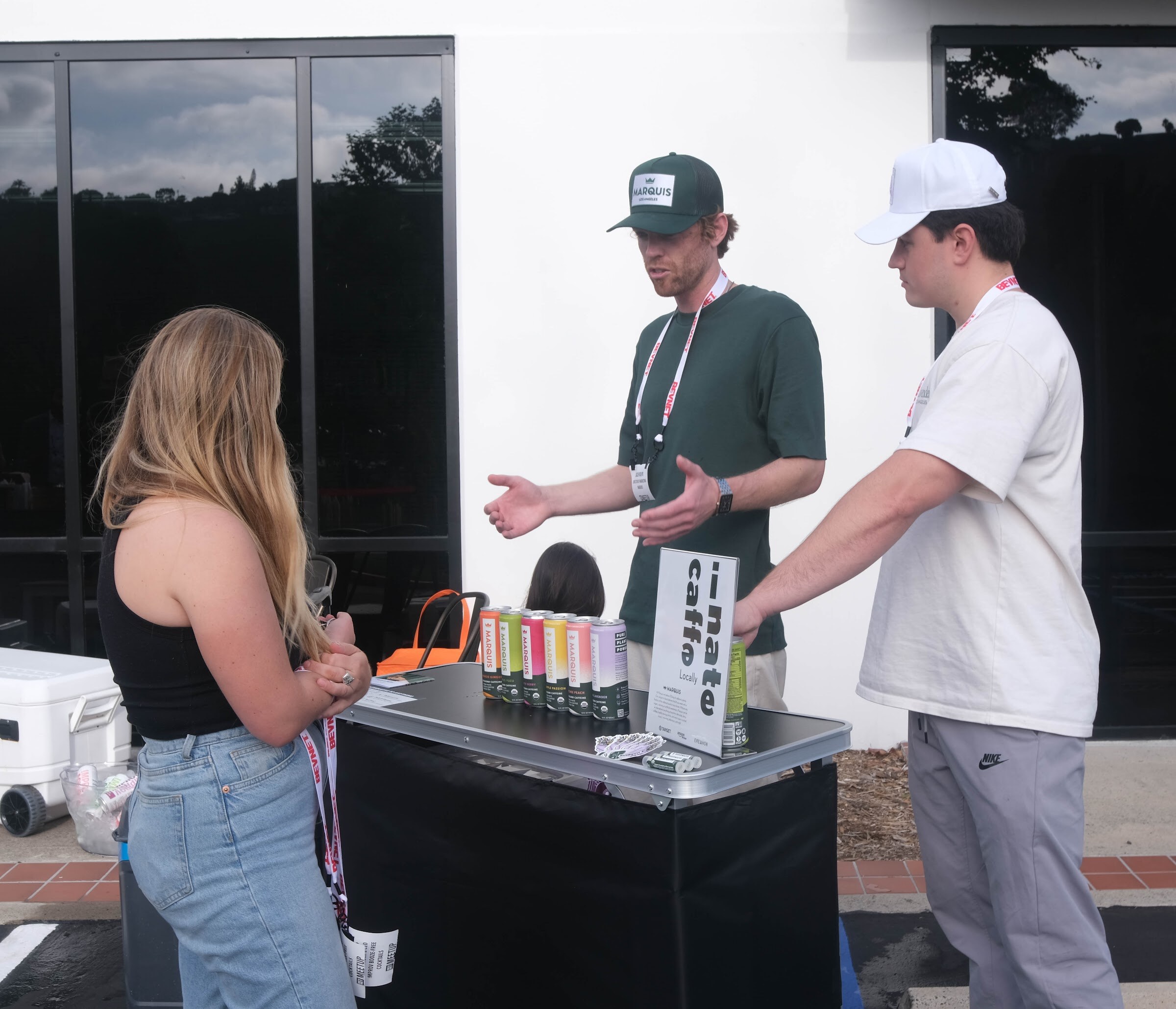 Three people in lanyards talk beside a black table with a row of colorful iNate caffe cans, in front of a white building.