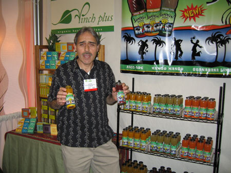 A man in a dark patterned shirt holds two bottles in front of shelves of bottled drinks, under a Vinch plus sign and a banner with palm tree graphics.