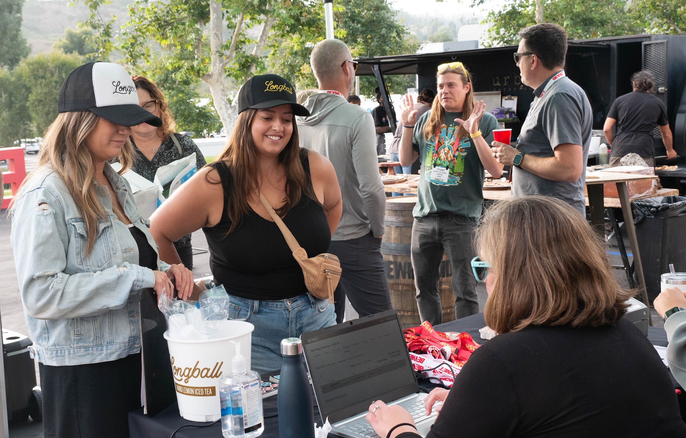 Women in caps talk with other guests at a table with a Longball bucket and a laptop.