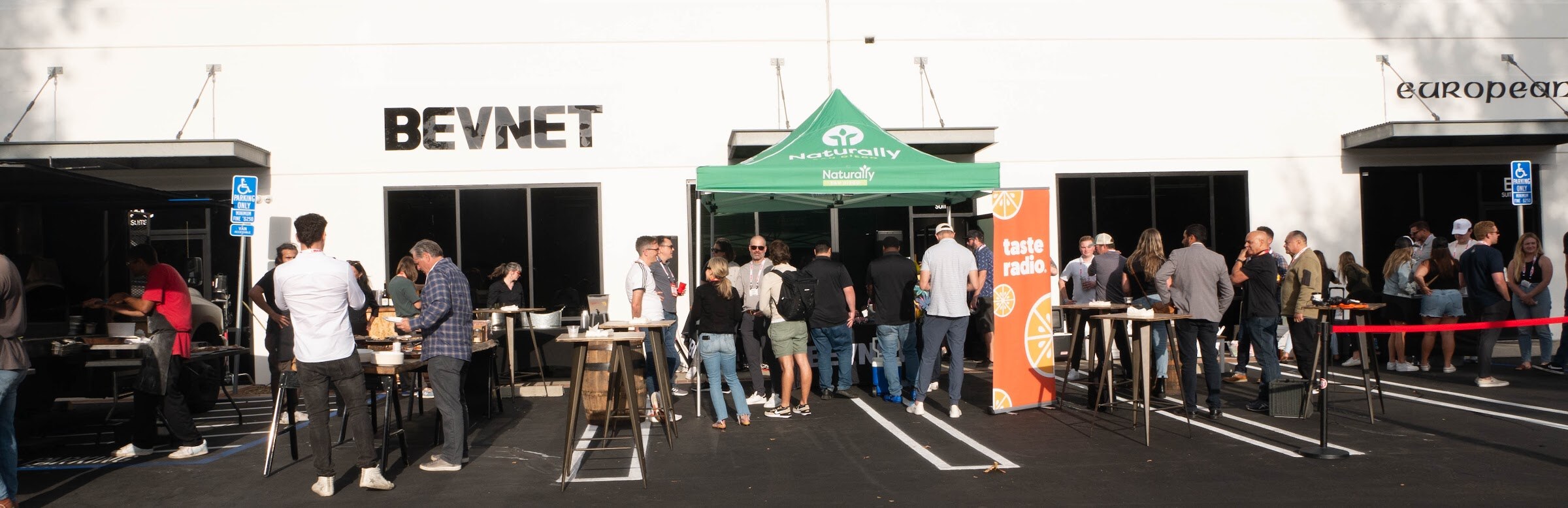 Wide view of guests standing in a parking lot in front of a white building with a BEVNET sign and a green Naturally tent.