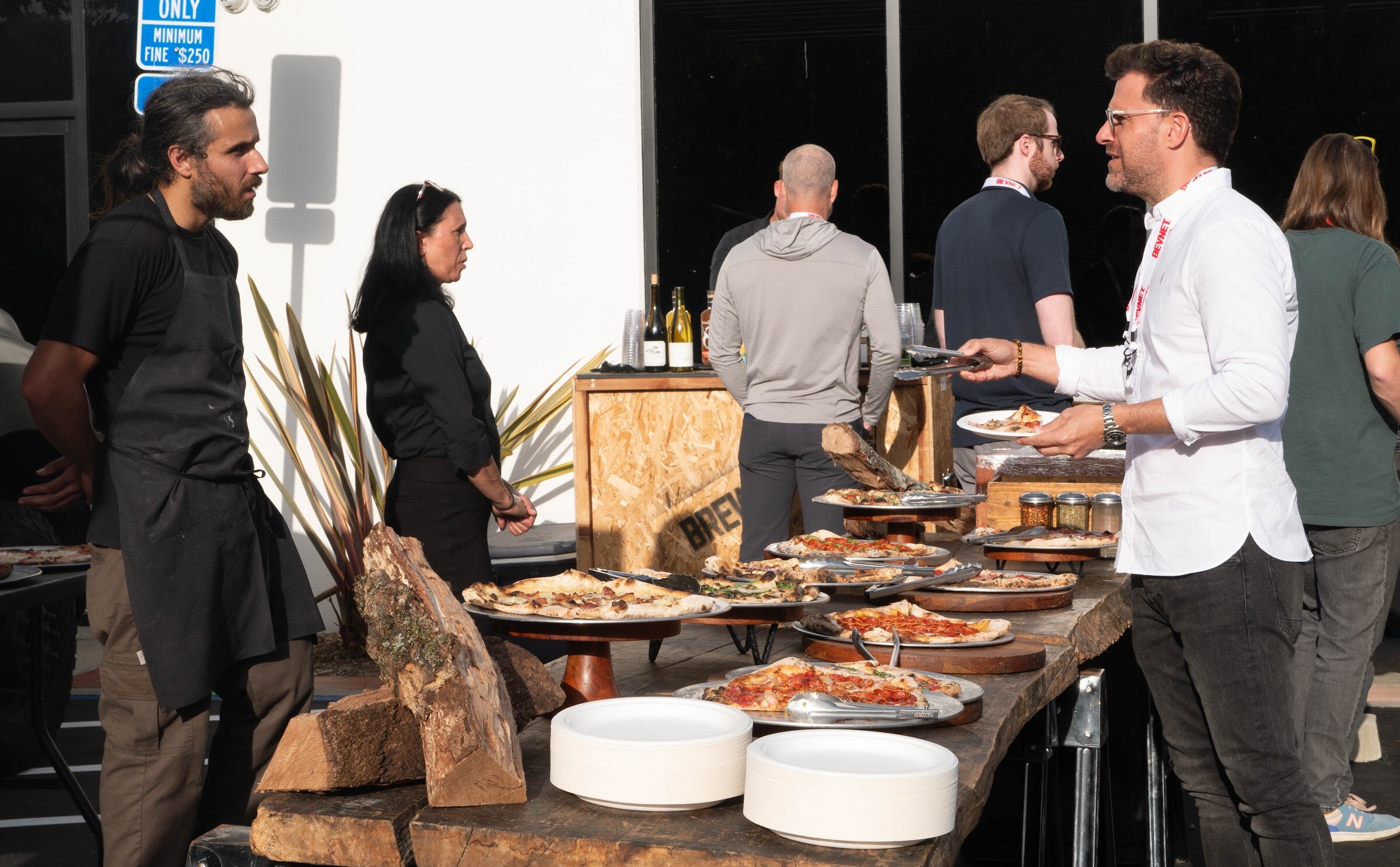 Guests serve themselves from a long wooden table of pizzas while a chef in black stands behind it.