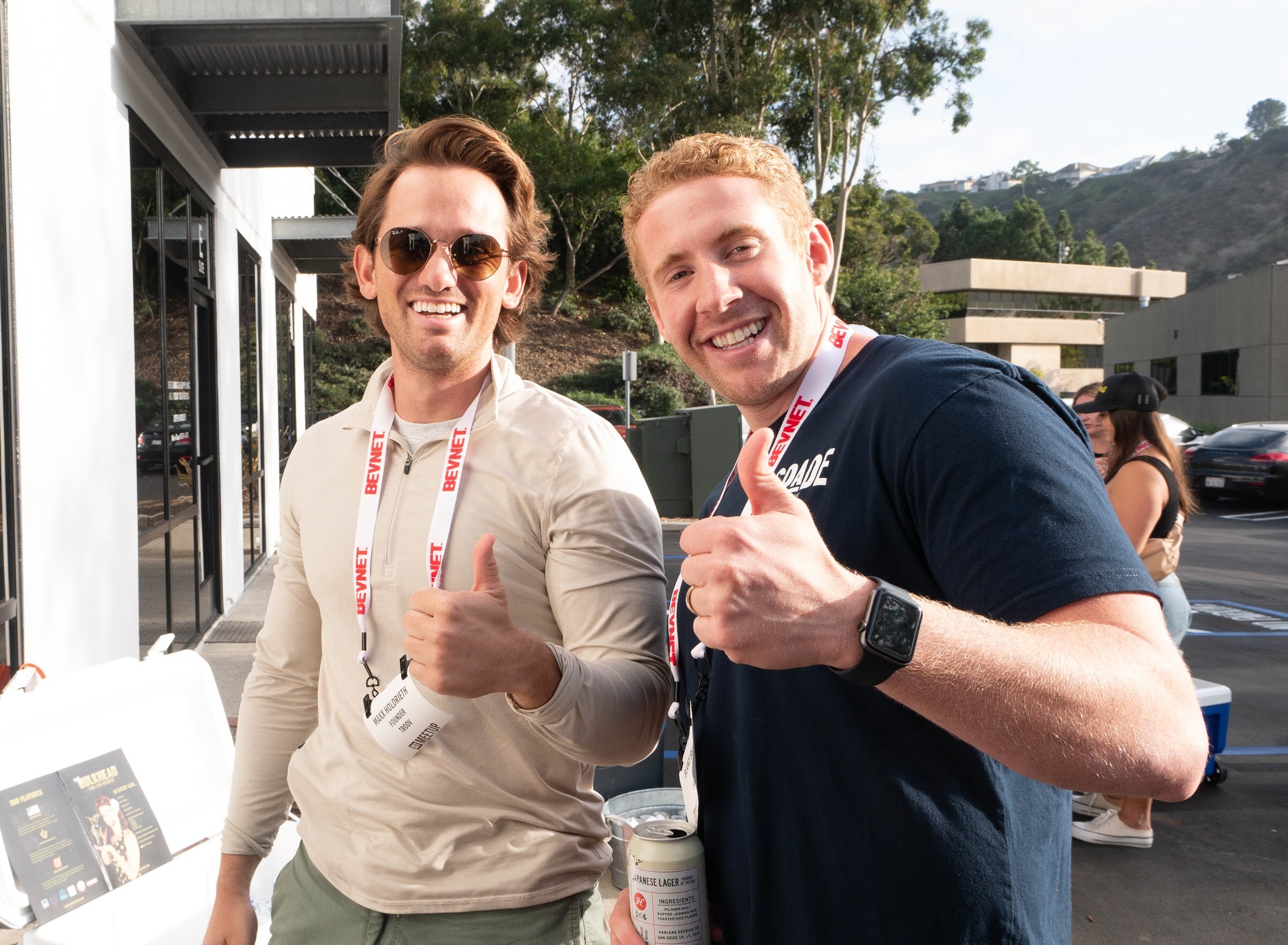 Two smiling men in lanyards, one in sunglasses and one in a navy shirt, give thumbs up in the sun.