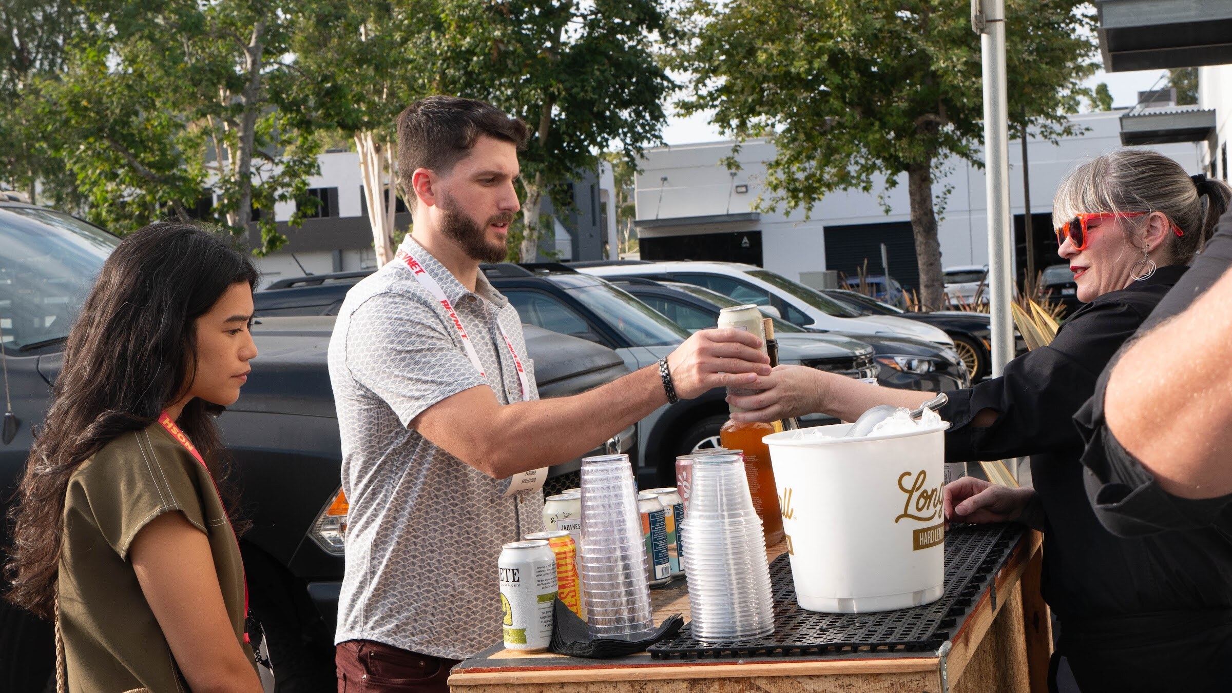 A bearded man hands a canned drink to a woman while a woman in sunglasses stands behind a Longball ice bucket.