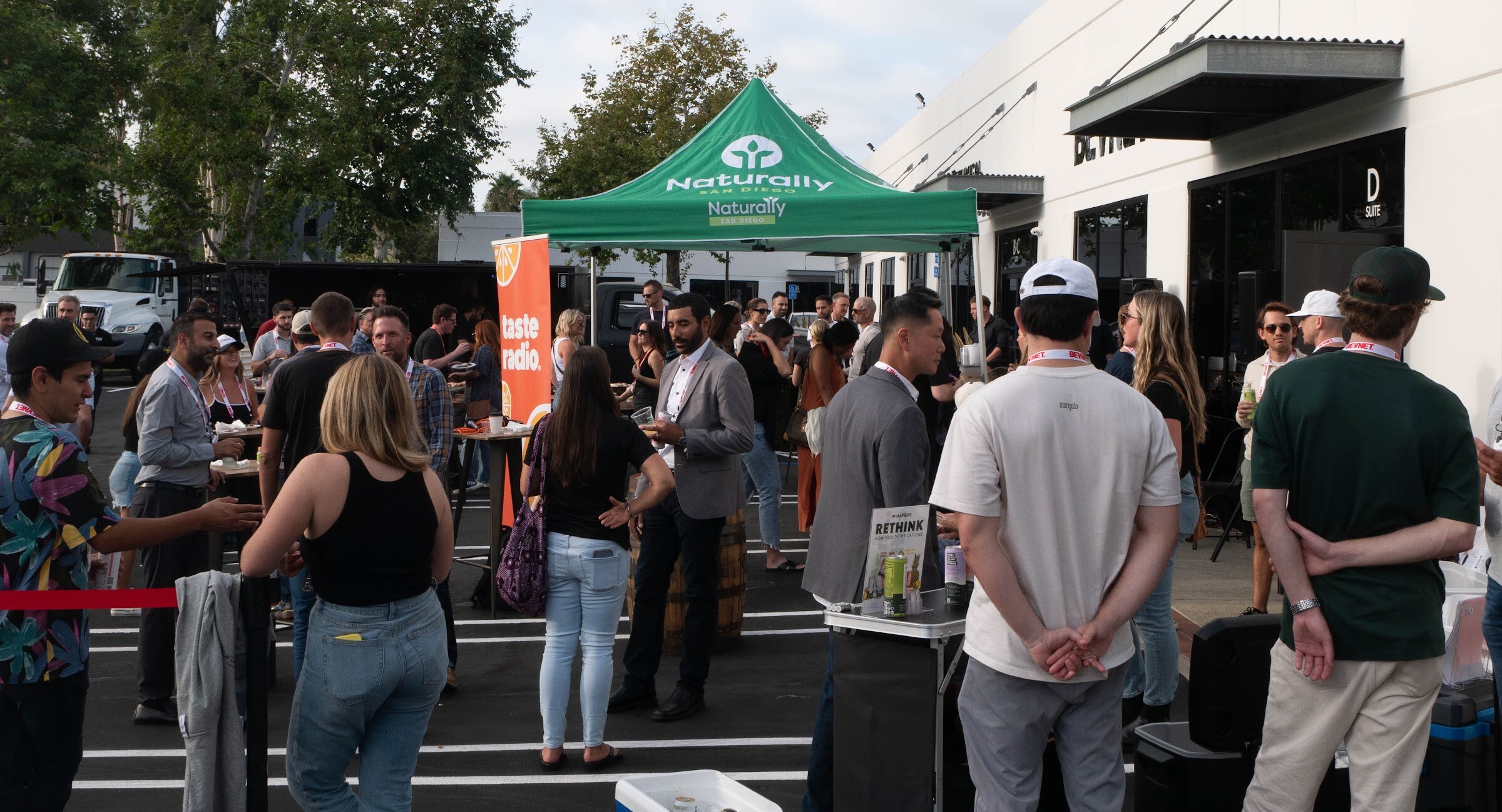 Guests mingle in a parking lot near a green Naturally tent and an orange banner.