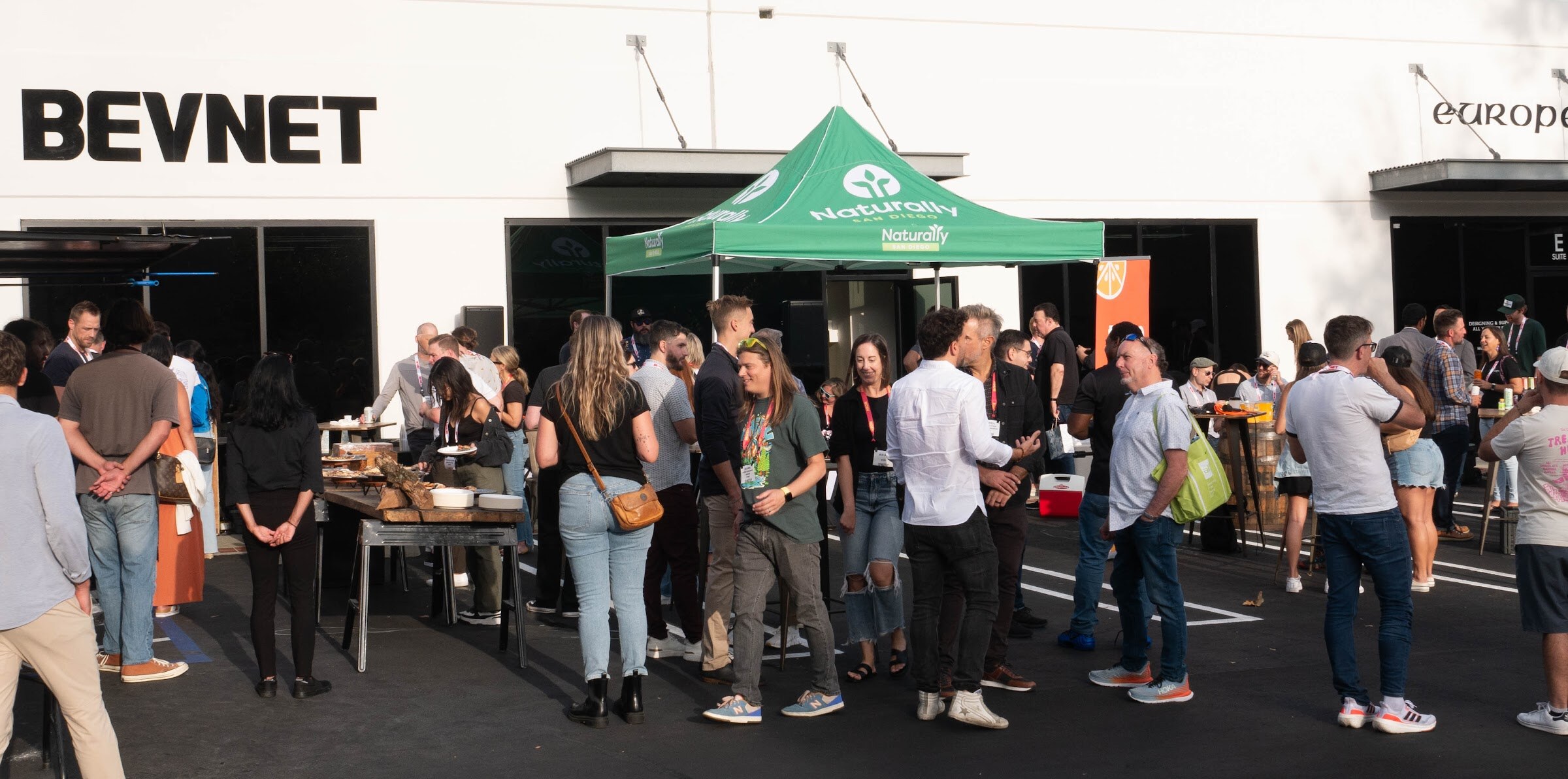 Wide view of guests standing at tables and under a green Naturally San Diego tent in front of the BevNET building.