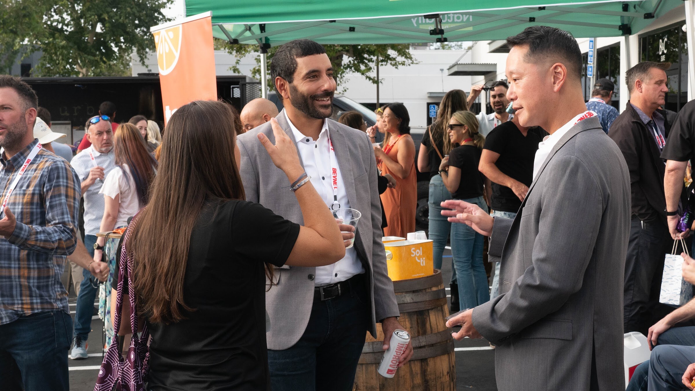 A man in a gray jacket talks with a woman in black near a barrel with orange boxes and a crowd behind them.