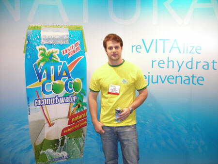 A man in a yellow-green shirt and jeans holds a small can beside a large Vita Coco coconut water carton graphic. A blue backdrop reads revitalize, rehydrate, rejuvenate.