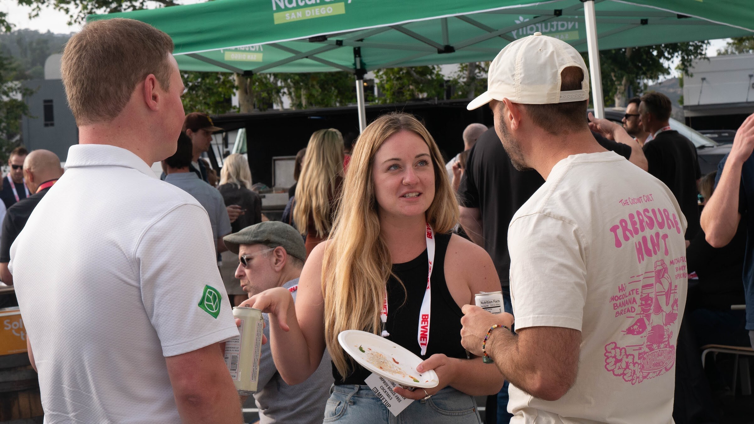 A blonde woman with a plate talks with two men in caps while others mingle under a green tent.