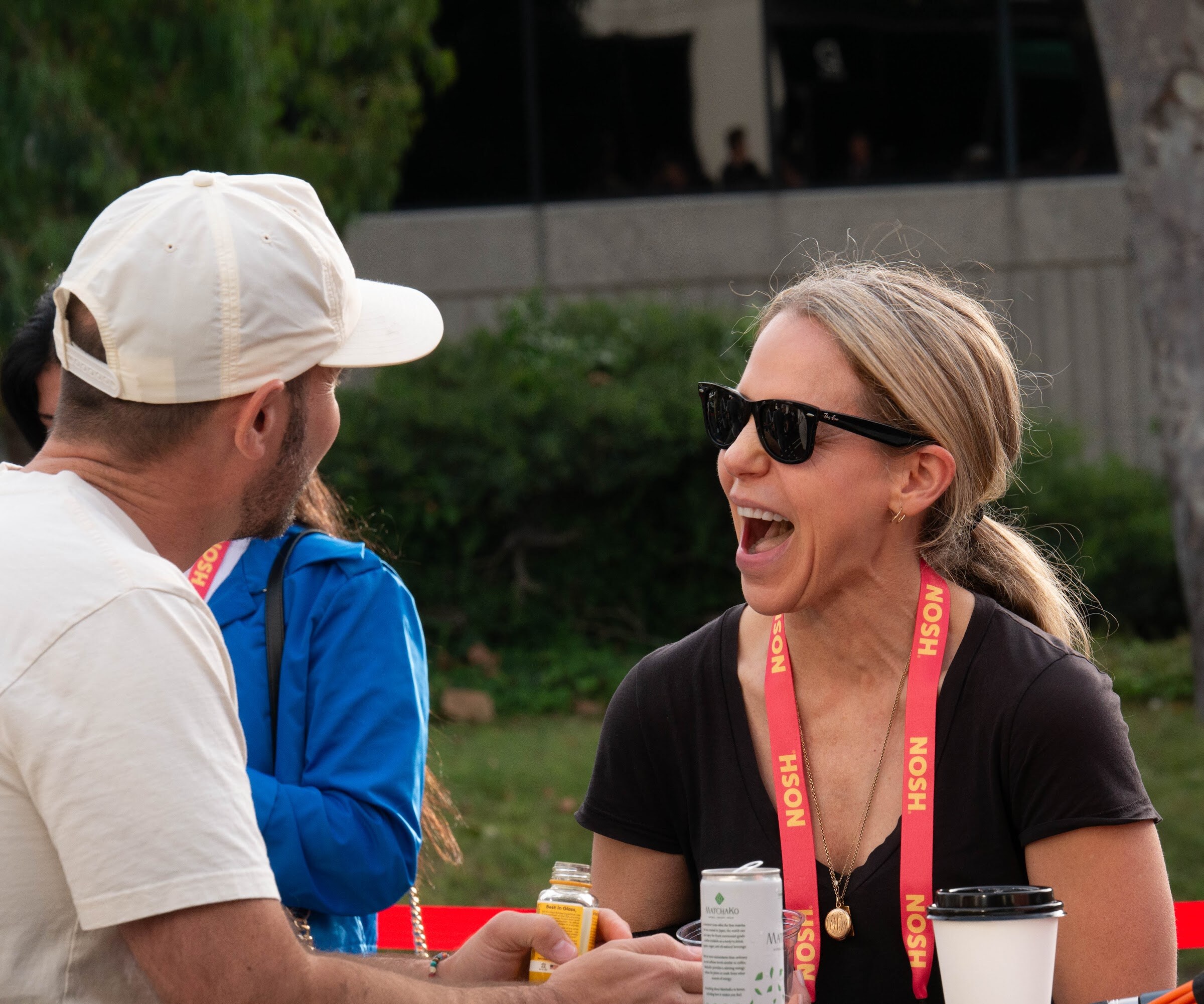 A woman in sunglasses laughs while a man in a cream cap talks to her, with grass and trees behind them.