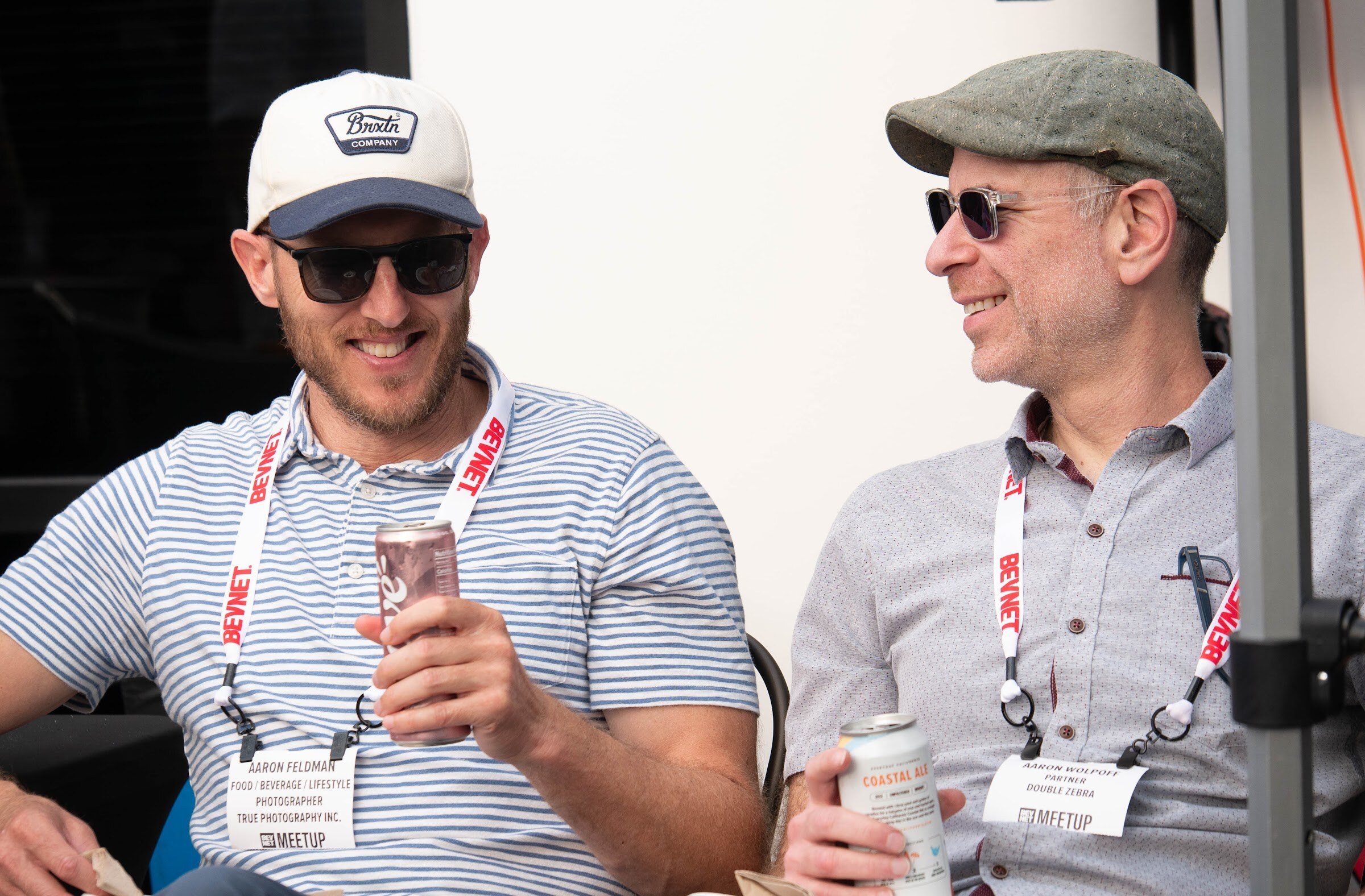 Two men in sunglasses and lanyards smile and hold cans at a table in a tent.