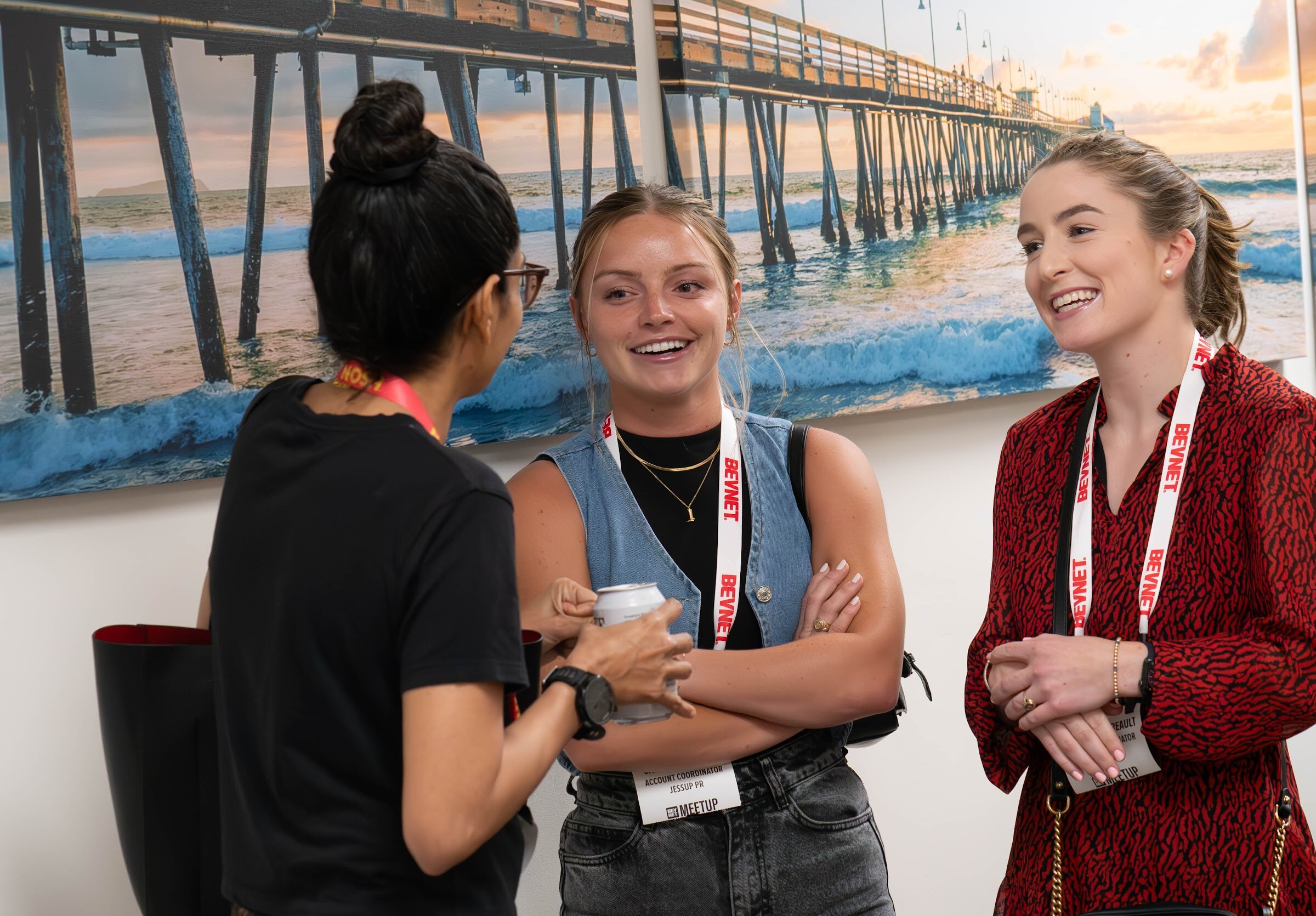 Three women with lanyards talk inside an office, with a large photo of a pier and sunset behind them.