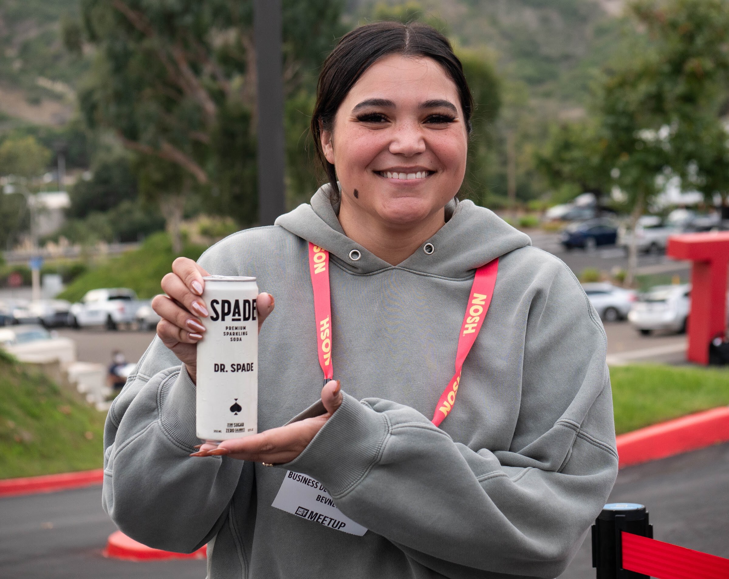 A smiling woman in a gray hoodie and Nosh lanyard holds up a can of Dr. Spade premium soda outdoors.