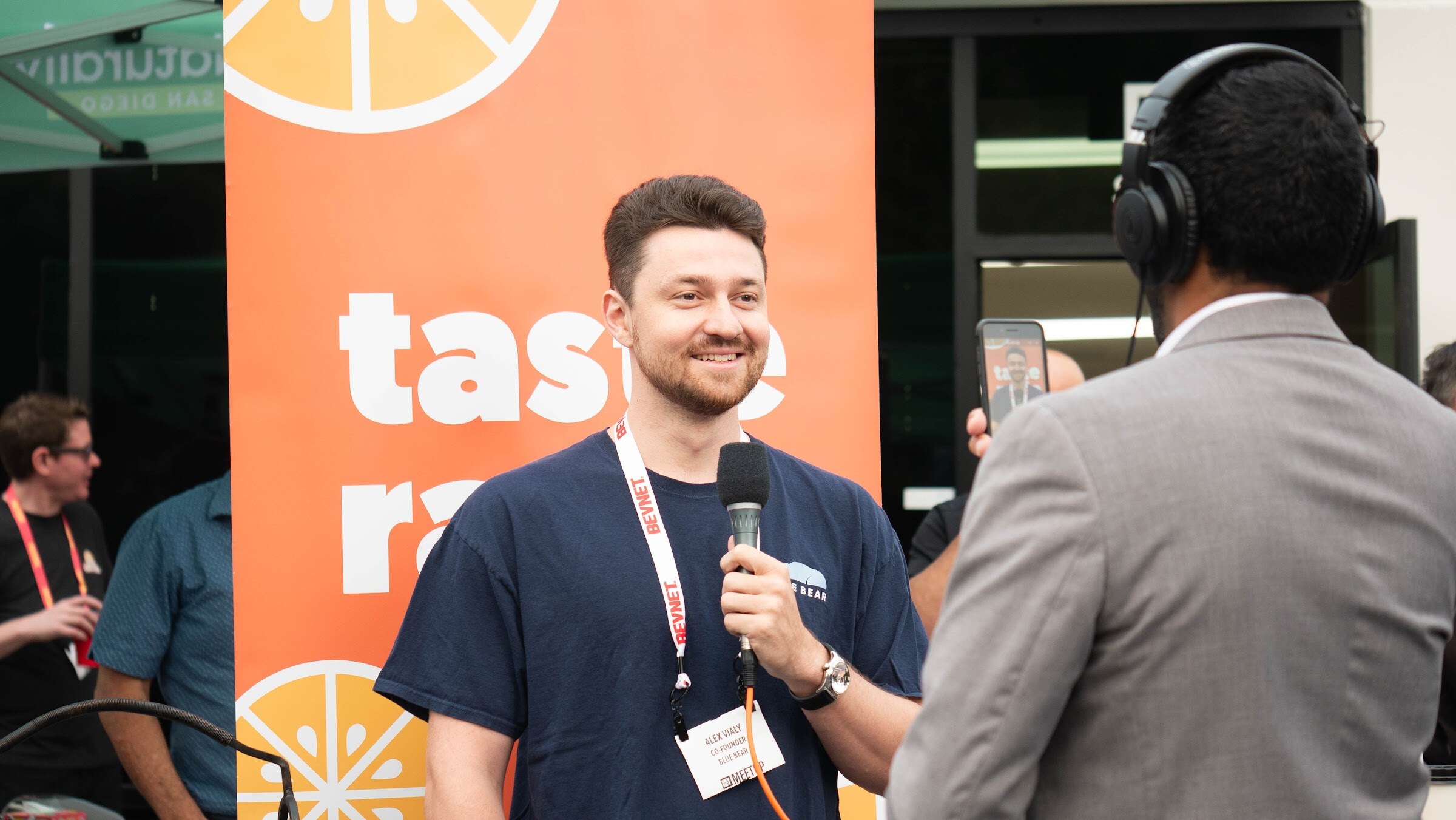 A smiling man in a blue shirt holds a microphone in front of an orange Taste Radio banner while a man in a headset and suit listens.