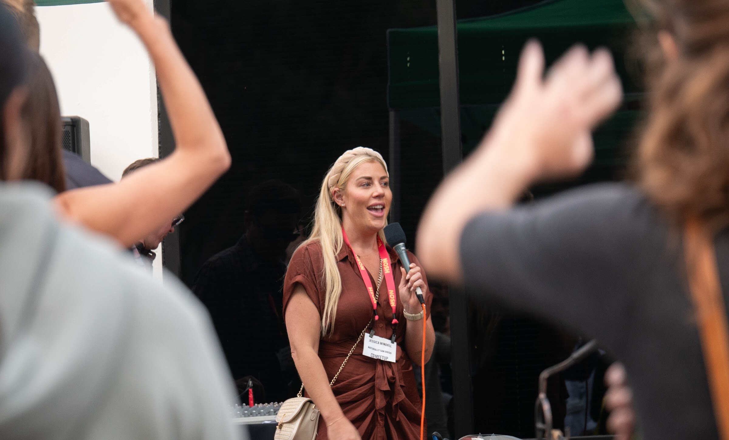 A woman in a brown dress speaks into a microphone while guests raise their hands in front of her.