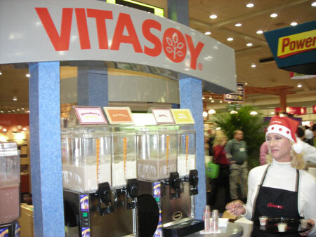 A Vitasoy sign hangs above a row of beverage dispensers while a woman in a red Santa-style hat and black apron holds small sample cups at the booth.