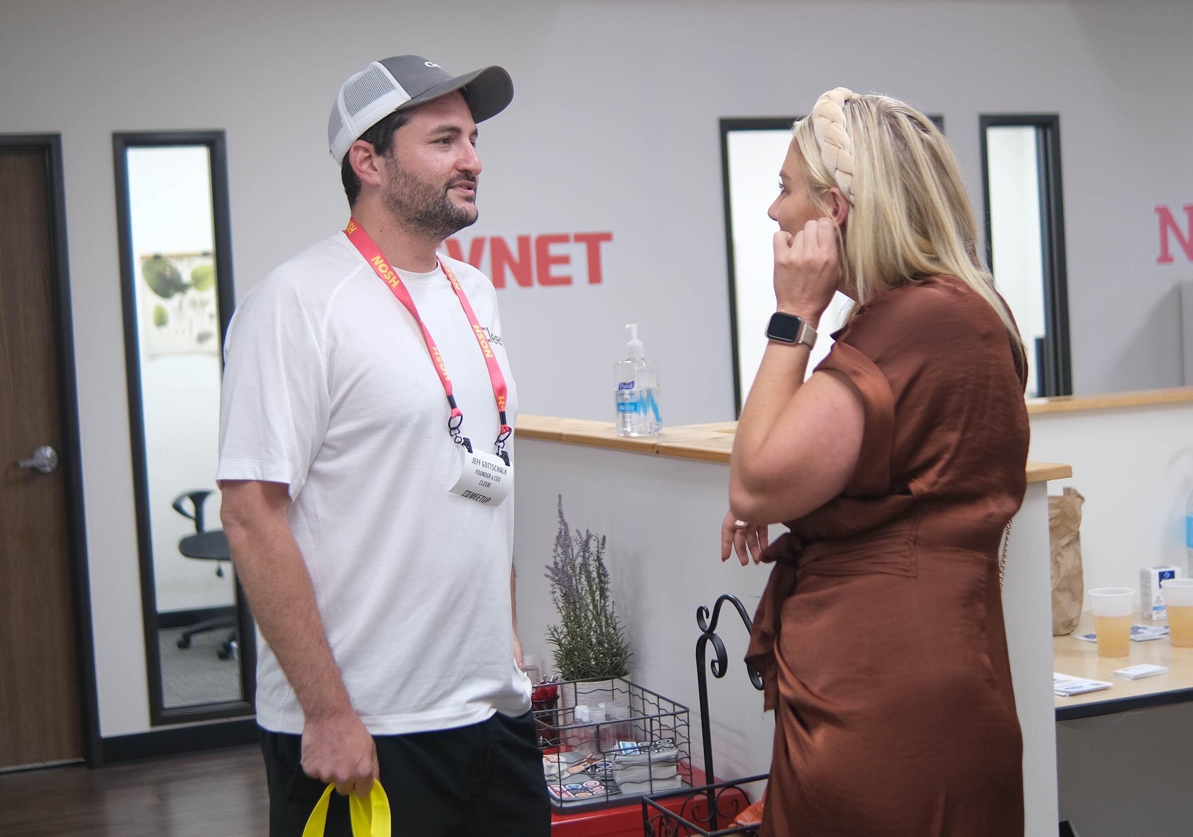 A man in a white shirt and a woman in a bronze dress talk near a counter in the office lobby with a BEVNET sign.