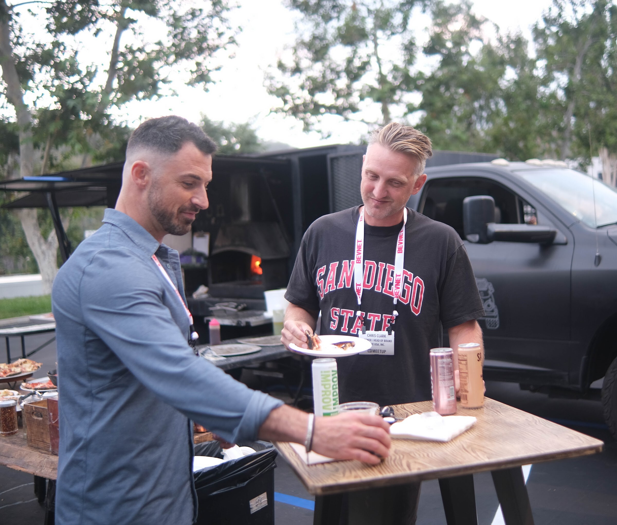 Two men in lanyards serve themselves at a wooden table outdoors with a pizza oven truck behind them.