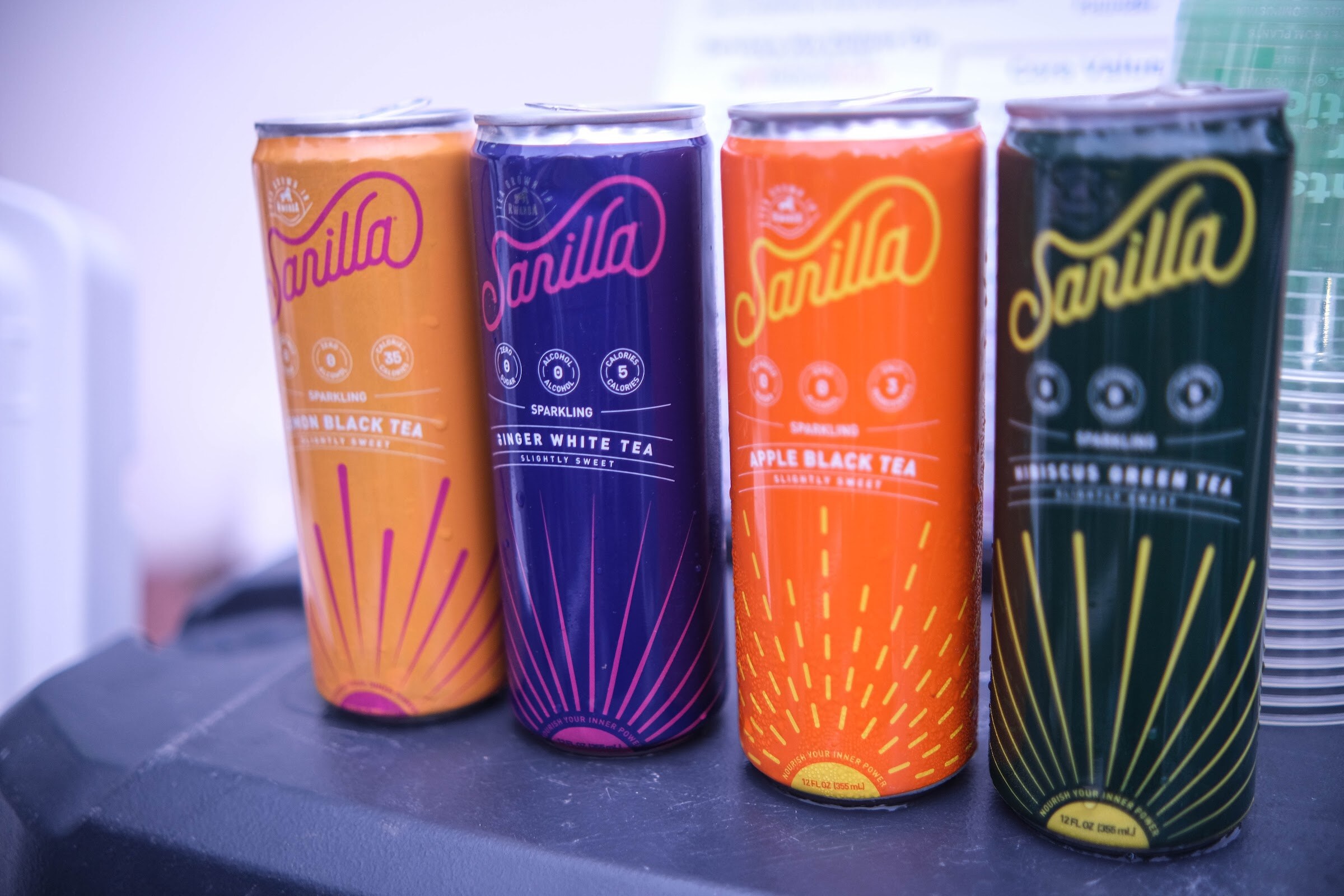 Four tall cans of Sanilla sparkling tea in orange, purple, red and dark green designs.