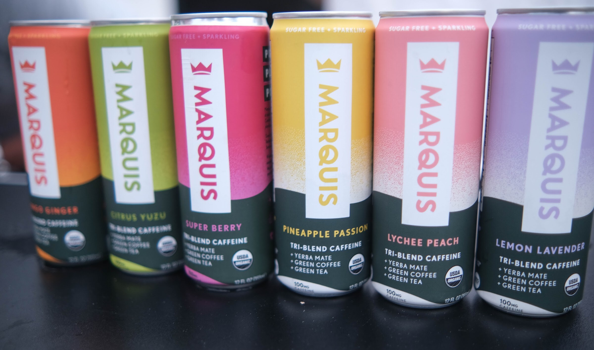 Six tall Marquis cans in assorted colors lined up in a row on a black table.